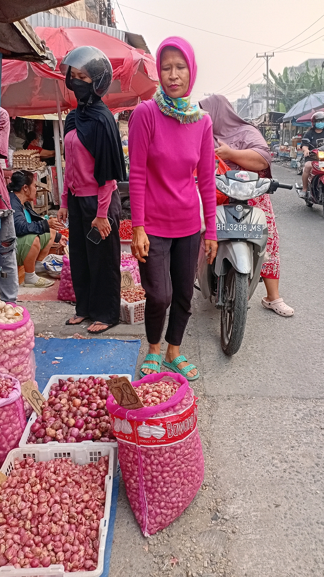 A woman wearing a pink hijab and a bright pink sweater stands on a street in Jambi, Indonesia, in front of a market stall. She is looking slightly to her left, with a neutral expression. Behind her, another woman, partially obscured, wears a black hijab and helmet, a pink sweater, and black pants. In the foreground, there are several large sacks and containers filled with small red onions, some displayed on a blue tarp on the ground. A motorcycle is parked to the right of the woman in the pink sweater. The street is paved, and the background shows more market stalls, buildings, and trees, suggesting a busy urban market environment. The sky is overcast.
