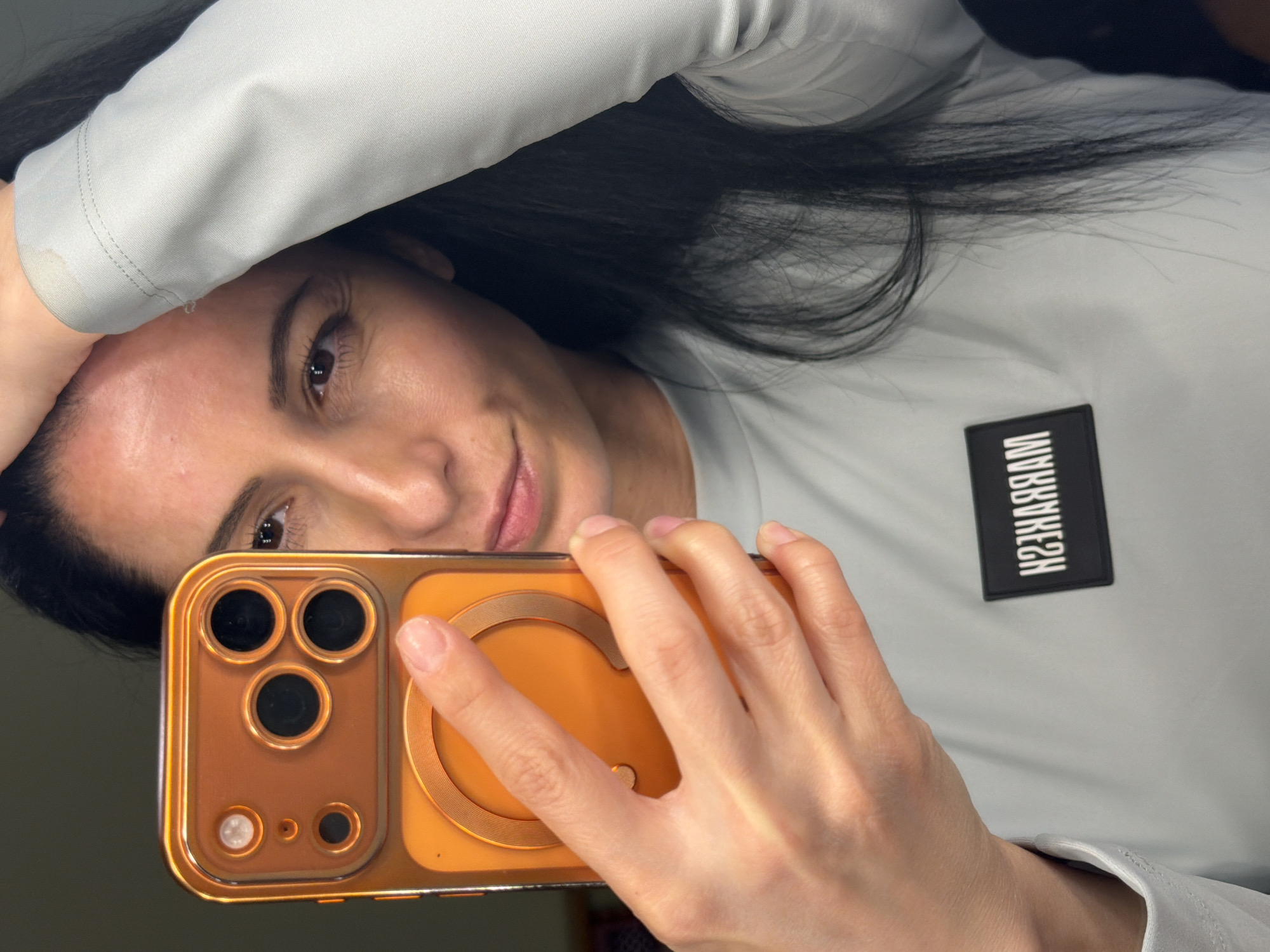 A selfie shows a woman with dark hair, brown eyes, and fair skin holding an orange-colored smartphone in front of her face. Her right hand is visible, gripping the phone, while her left hand rests on her head. She wears a light gray, long-sleeved shirt. A small, black rectangular patch with white text that reads "H23ЯЯАЯАМ" is sewn on the right side of her shirt. The background is nondescript. The photo was likely not taken in Pleven, Bulgaria, as there are no elements identifiable to the area in the photograph.