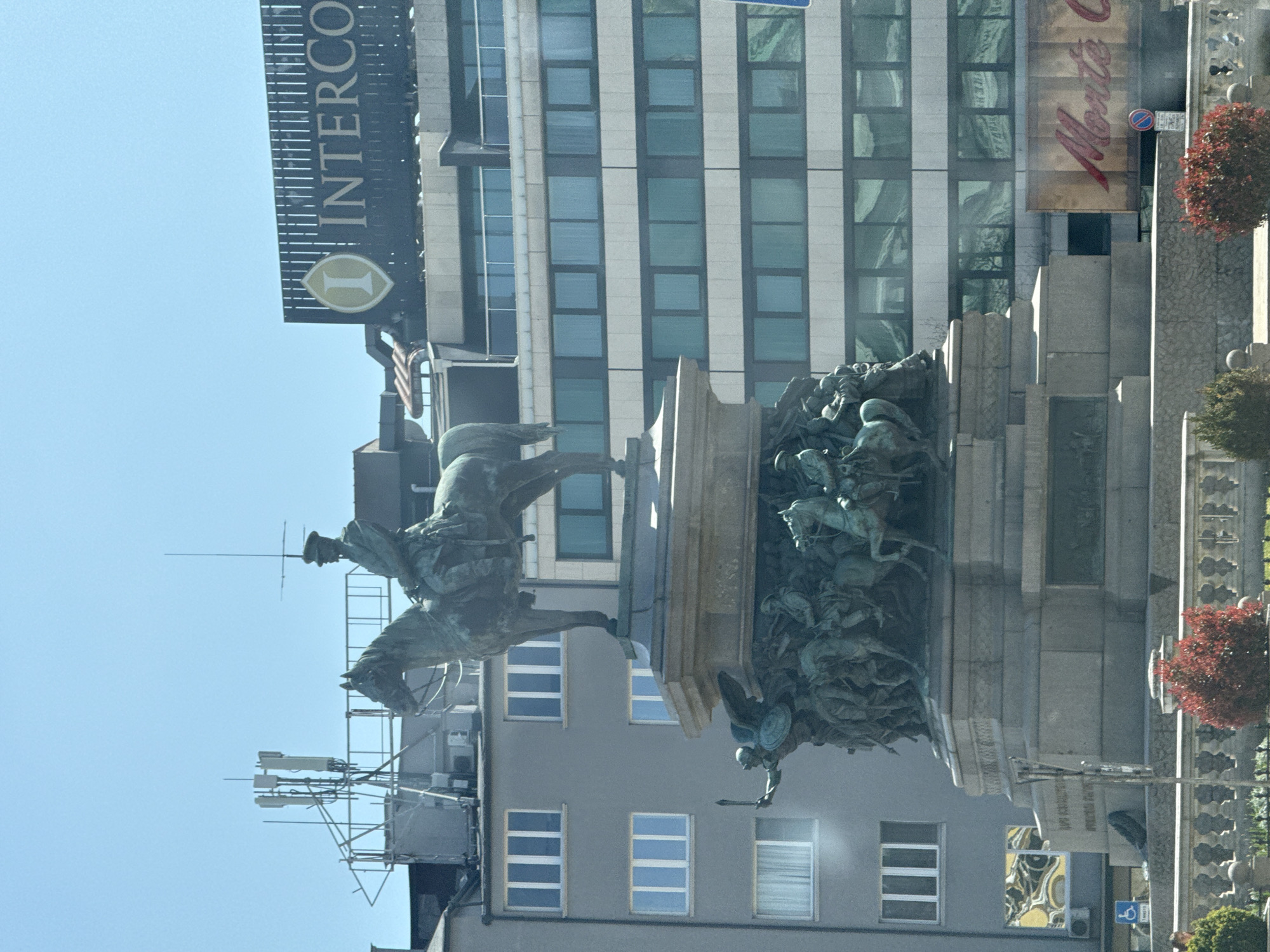 The image depicts the equestrian statue of Prince Michael in Republic Square, Belgrade, Serbia. The statue stands prominently in the square, with an InterContinental hotel and other buildings visible in the background. The statue itself features Prince Michael on horseback, with additional bronze figures at the base representing important historical figures. The surrounding environment includes trees and street furniture.