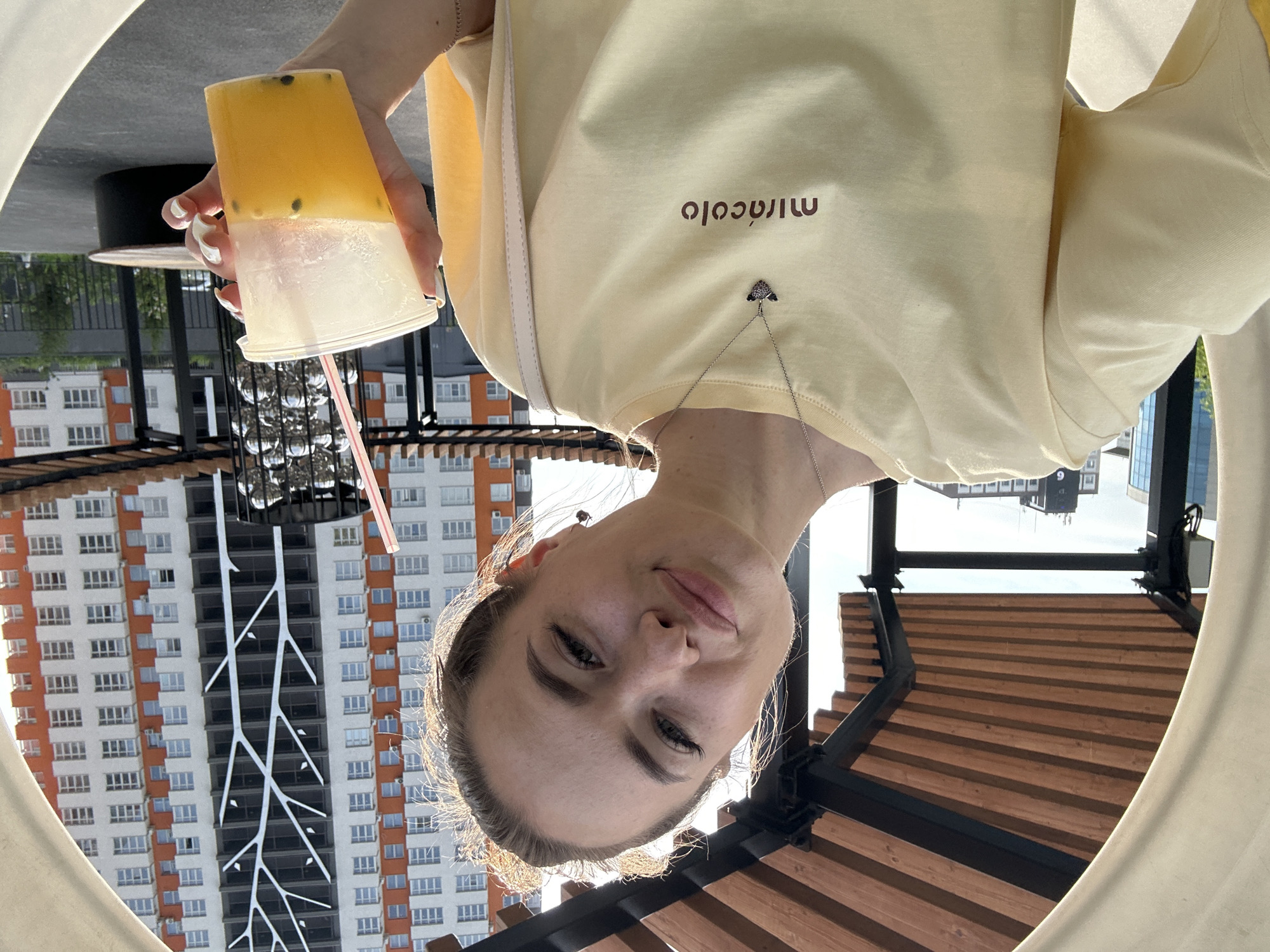 A young woman with her hair pulled up is taking a selfie. She is wearing a pale yellow t-shirt with the word "miracolo" printed on it in brown lettering. She has a silver necklace with a small charm. She is holding a clear plastic cup with an orange beverage and a pink straw. In the background, there are several modern buildings, one of which has a white tree-like design on its facade. There is also a wooden structure overhead. The scene appears to be outdoors in an urban setting during the daytime.