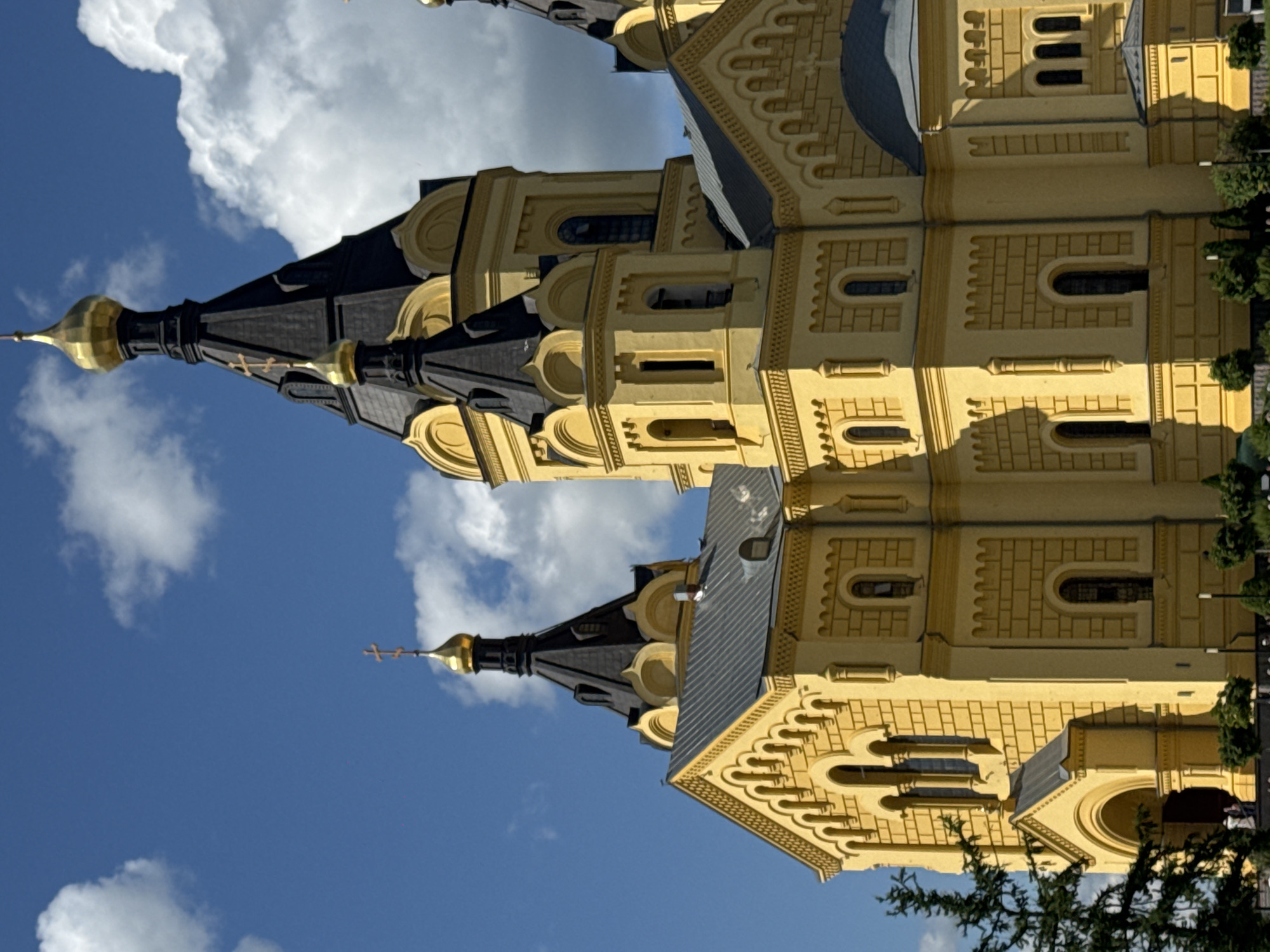 The image shows the Alexander Nevsky Cathedral, a prominent Orthodox cathedral located in Nizhny Novgorod, Russia. The cathedral is painted a vibrant yellow and features a dark, multi-tiered roof topped with golden domes. Multiple smaller golden domes are visible on the spires, some adorned with crosses. The architecture exhibits intricate detailing, including arched windows and decorative brickwork patterns. The sky is a bright blue with scattered white clouds, suggesting a sunny day. In the foreground, a dark green coniferous tree partially obscures the left side of the cathedral. The base of the cathedral is surrounded by green bushes and trees, and there are a few indistinct figures of people near the entrance on the lower left. The scene is captured from a slightly low angle, emphasizing the height and grandeur of the cathedral.