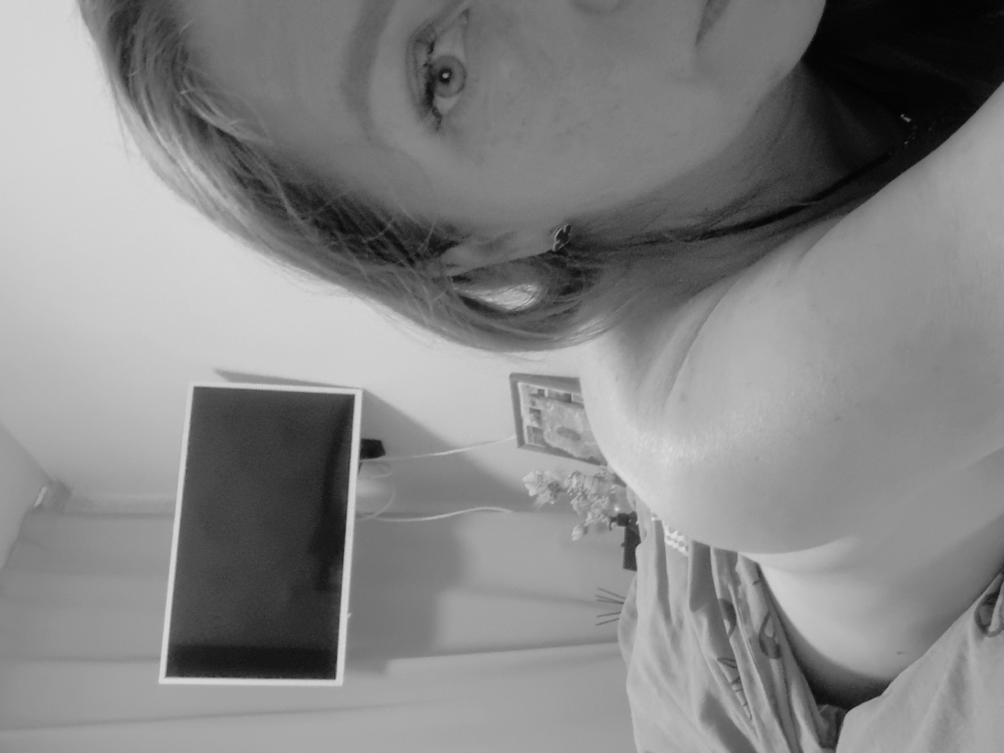 A grayscale image depicts a person from a low-angle perspective. The person has fair skin, visible freckles, and light-colored hair framing their face. An earring is visible in one ear. The person is wearing what appears to be a patterned garment. In the background, a framed rectangular object, likely a screen or picture frame, hangs on a wall. Adjacent to it is a framed picture and a vase containing flowers. The location and city of Chaikovsky, Russia cannot be independently verified within the image's content.