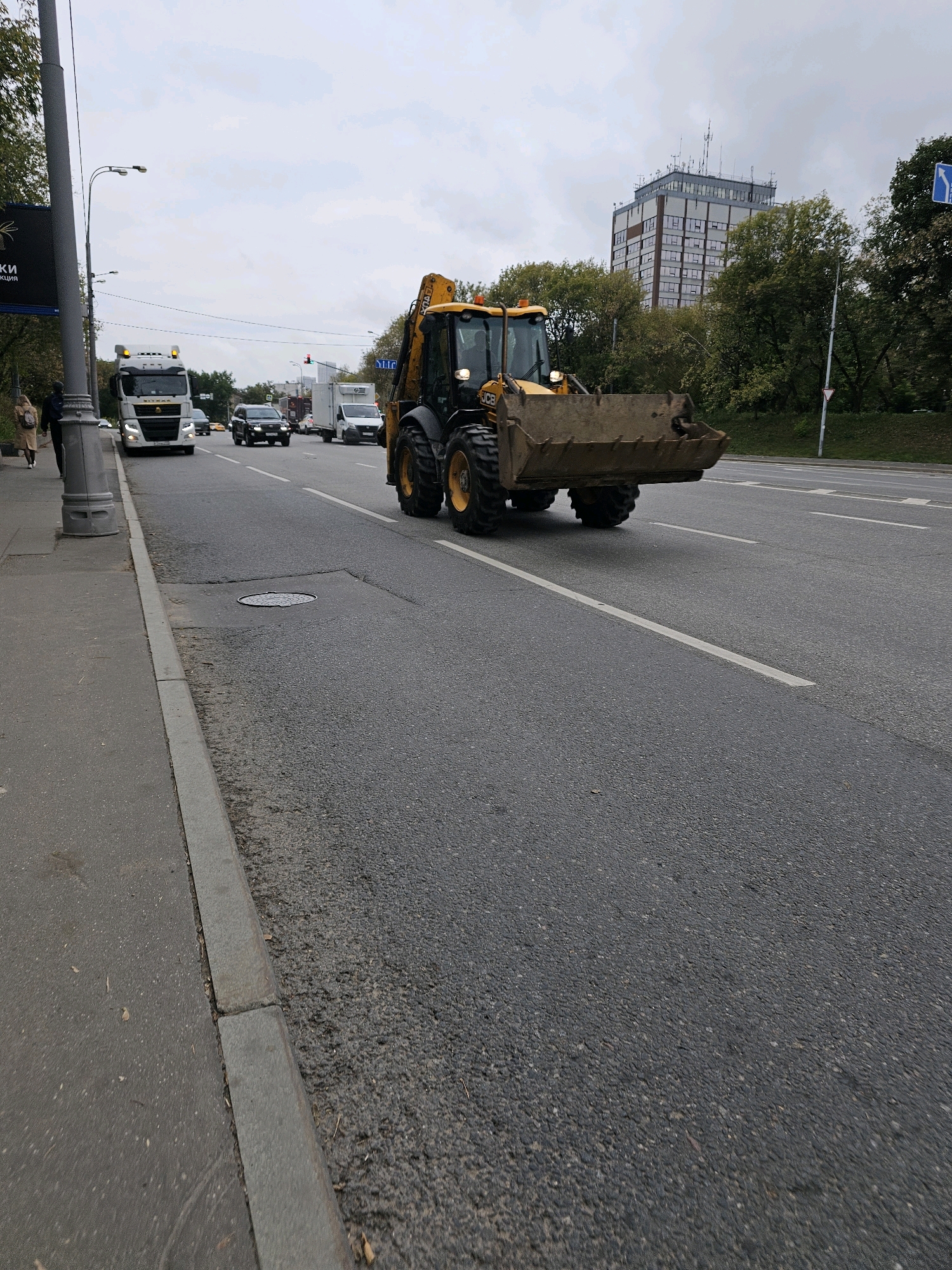 An overcast day in Moscow, Russia, shows a busy street scene. On the right side of the image, a yellow backhoe loader with its bucket raised is moving forward on the asphalt road. The machine has large, rugged tires. In the background, behind the backhoe, a tall, multi-story building with numerous windows and a visible antenna array on its roof stands amidst a grove of green trees. To the left of the backhoe, other vehicles are visible on the road, including a white truck with a trailer and a dark-colored SUV. On the far left, a sidewalk runs parallel to the road, bordered by a gray curb. A single street lamp is present on a pole. A few pedestrians are seen walking on the sidewalk. The road has white lane markings.