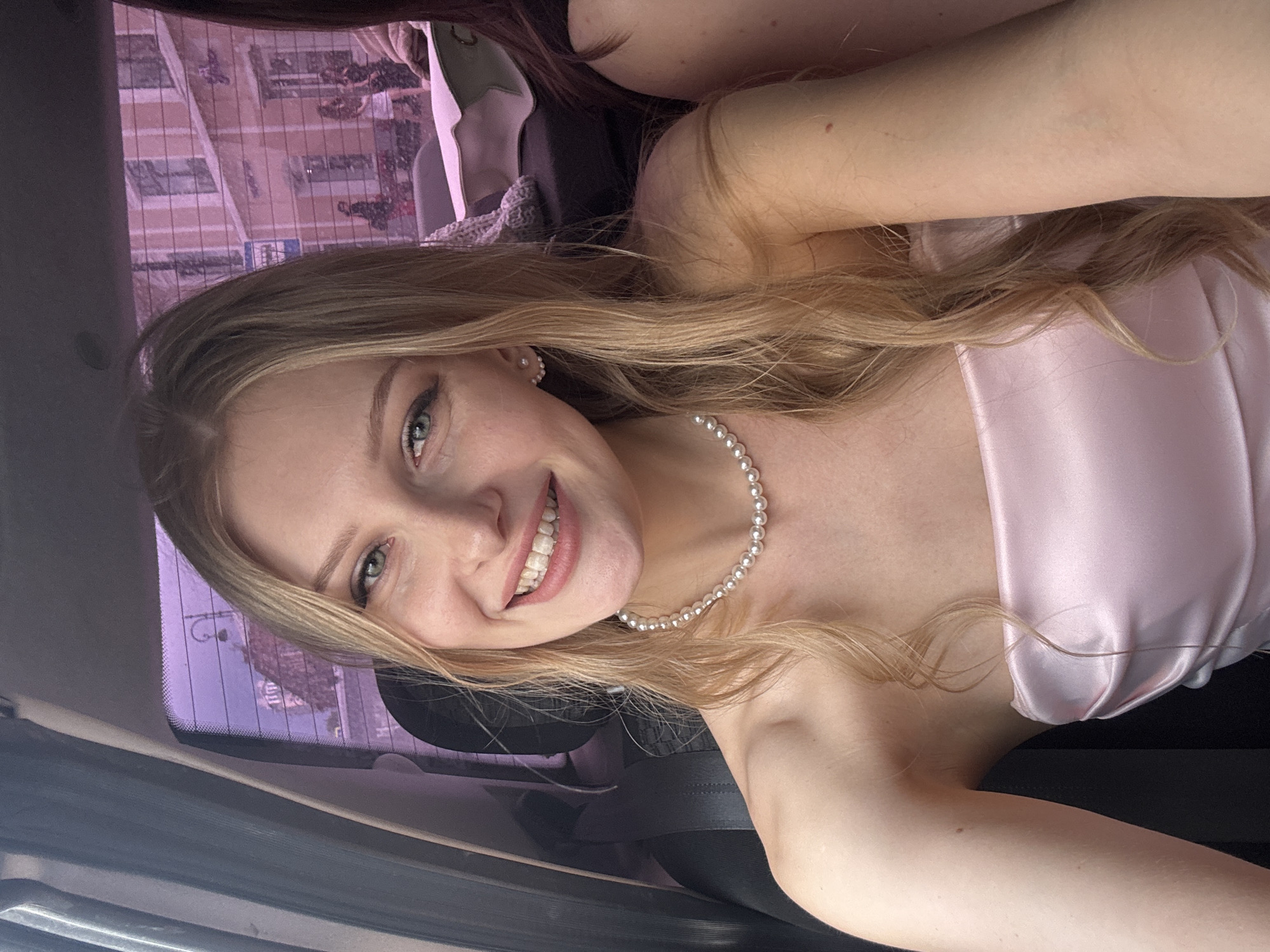 A woman is seated inside a vehicle, possibly a car, in Perm, Russia. The reflection in the car window suggests the urban environment of Perm. She is fair-skinned, with blonde hair, and is wearing a pearl necklace and a light pink, satin-like garment. She is smiling and looking directly at the camera. The interior of the vehicle is visible, and another person's hair can be seen at the top edge of the frame.