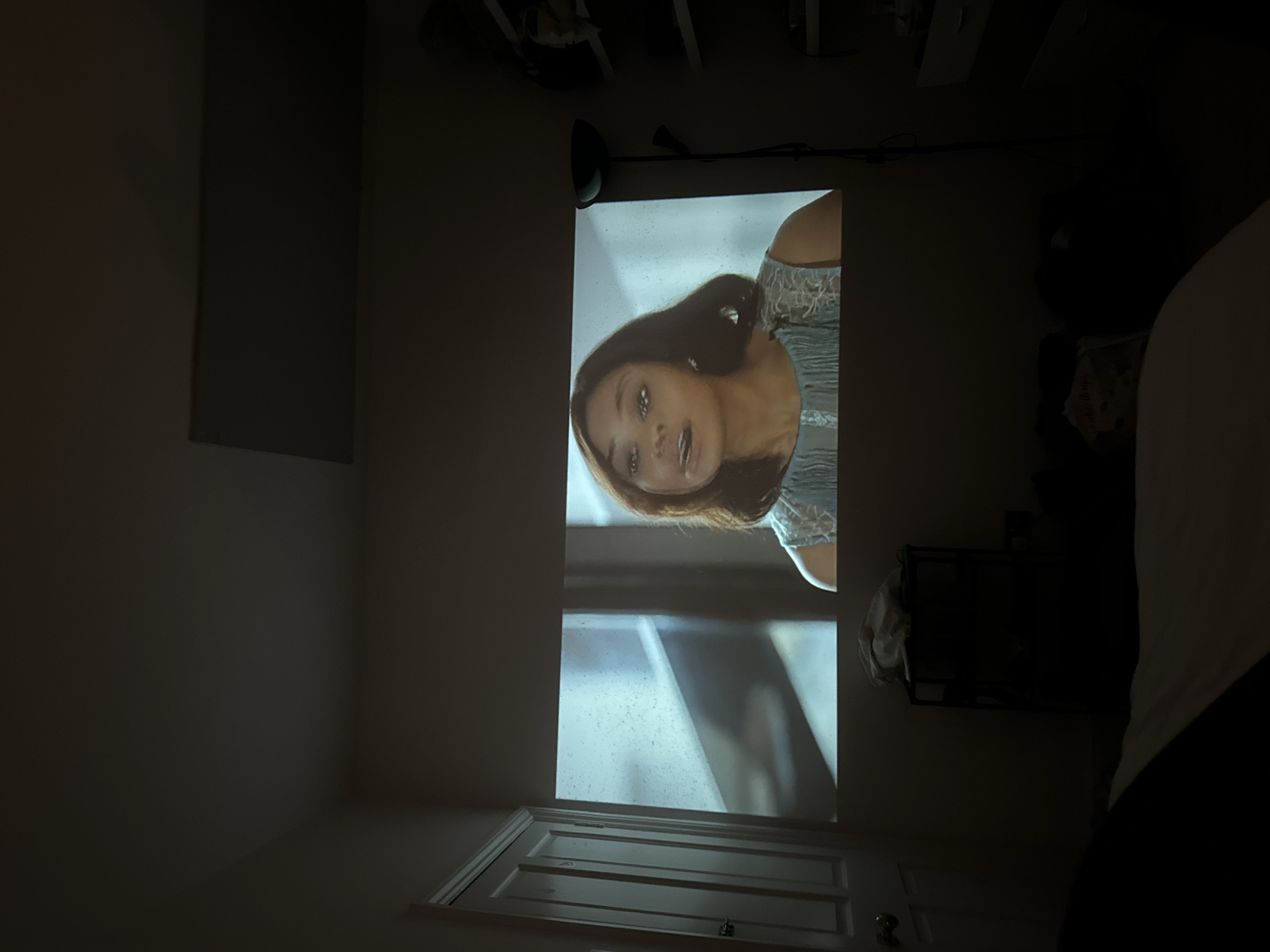 In a dimly lit bedroom, a projector displays an image of a woman with brown hair and fair skin, wearing a light blue dress. The woman is looking slightly to her left with her mouth open, as if speaking. The background of the projection shows a window with raindrops. To the left of the projected image is a white door with a doorknob. In front of the projection, a black shelving unit holds a white bag. A floor lamp stands to the right of the projection, and shelves with various items are visible on the right wall. The overall impression is of a relaxed evening at home, possibly watching a movie. The scene is devoid of any information that could place it in London, United Kingdom.