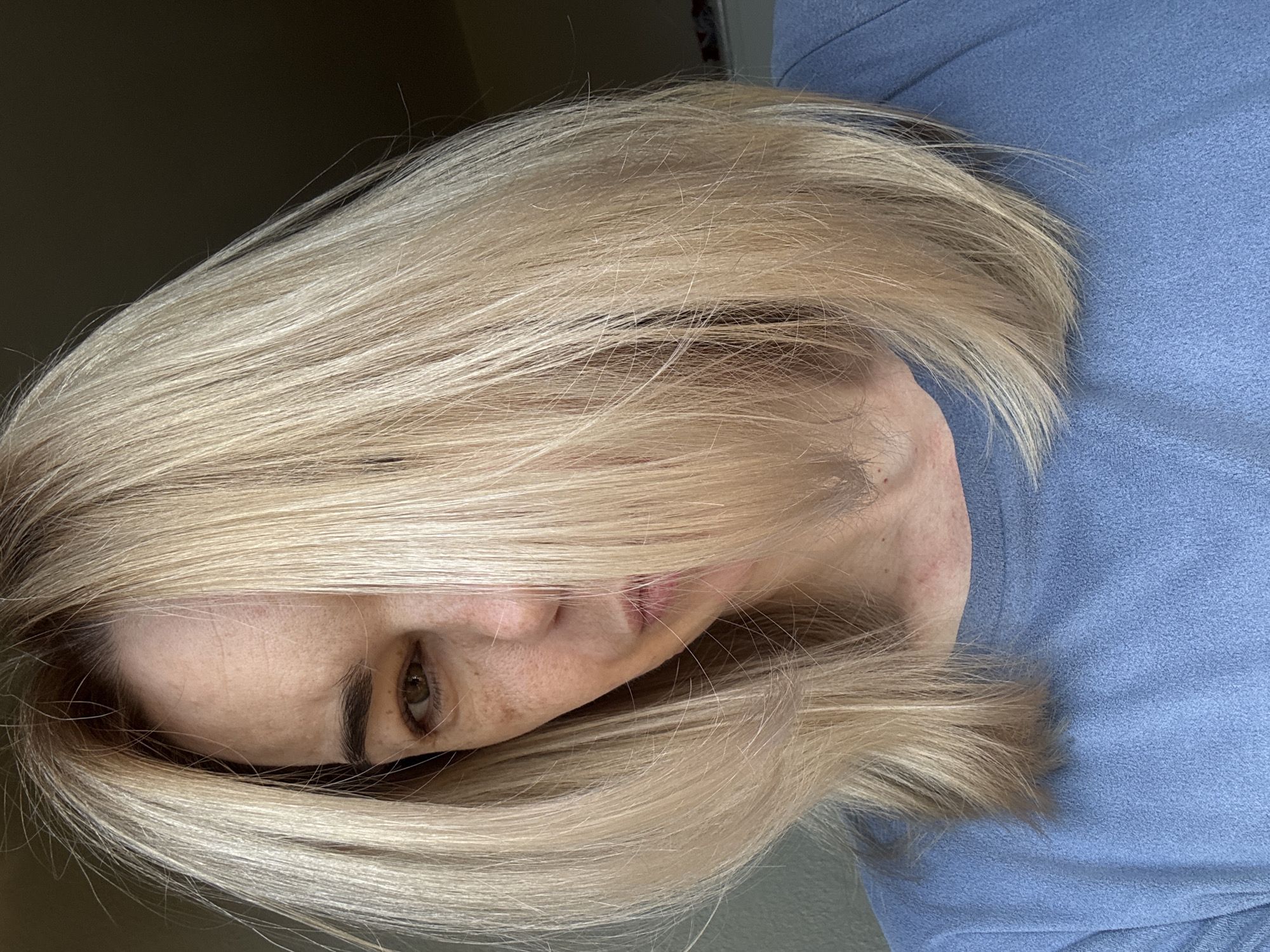 A woman with blonde hair is the central subject of the image. Part of her face is visible, including an eye, cheek, and lips. She is wearing a blue top. The background is out of focus, showing a light gray and dark brown wall. The image is taken indoors.