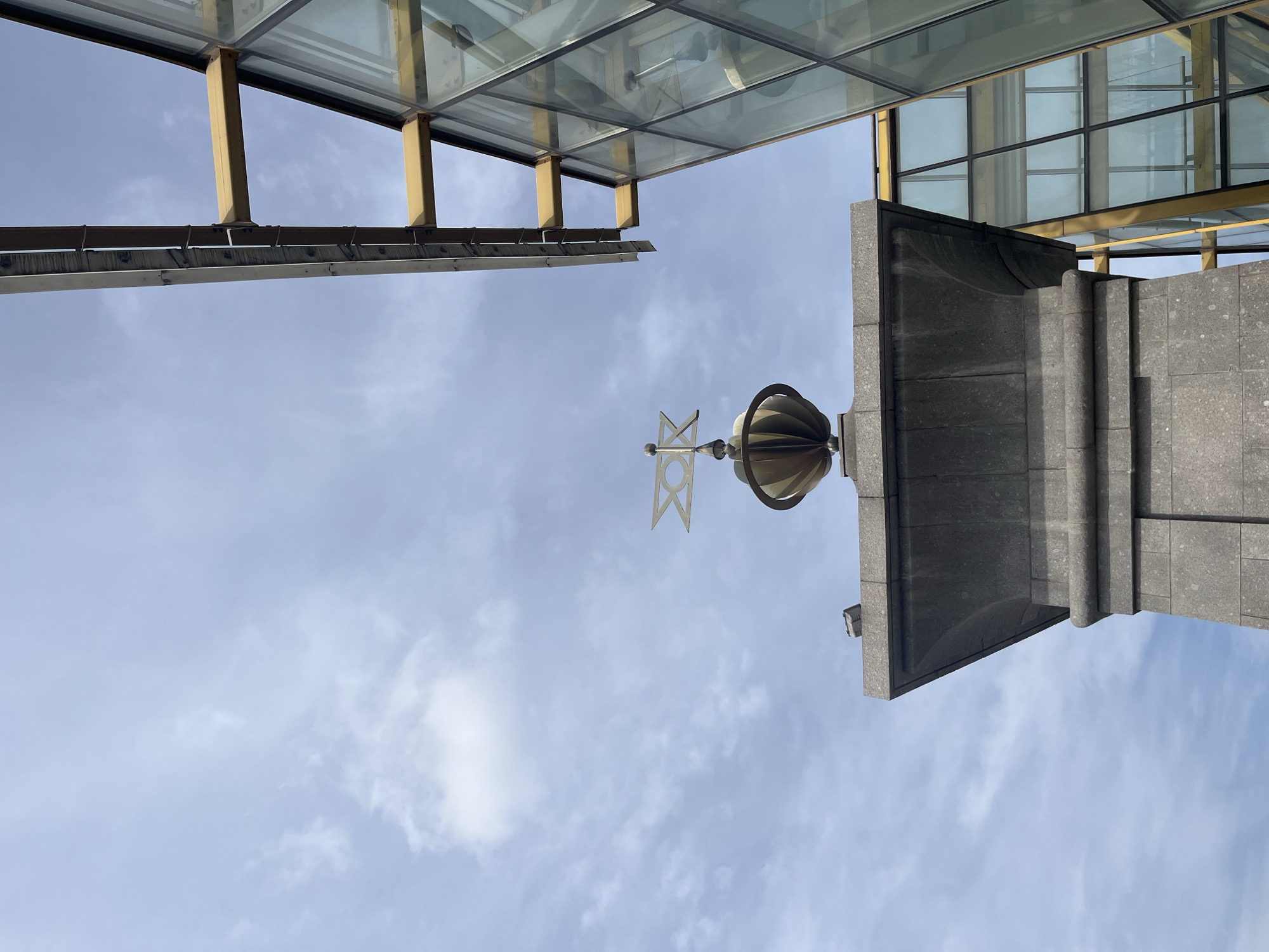 The photograph is taken from a low angle looking up at architectural elements against a light blue sky with scattered clouds.
A gray, architectural feature, possibly part of a building's facade or monument, is prominently displayed on the right. Atop this feature is a weather vane and a decorative orb.
Above and to the left, parts of a glass and metal structure are visible, perhaps a modern building or an overhead covering. A ladder extends toward the structure.
The location is Moscow, Russia.