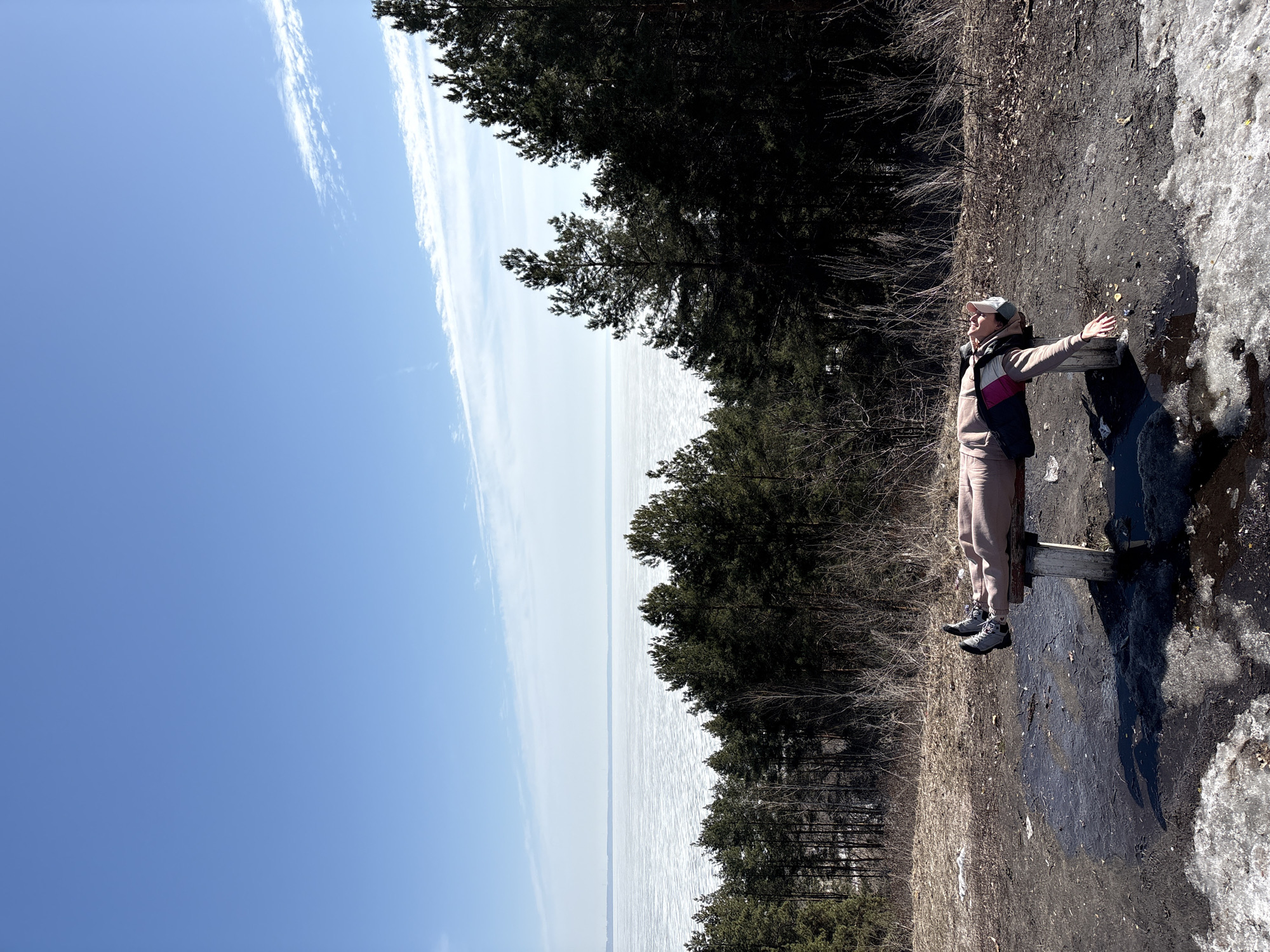A woman in a beige sweatsuit, vest, and cap sits on a wooden bench with her arms outstretched. The bench is located on a dirt and snow-covered bank overlooking a body of water, which appears to be a frozen river or lake. A line of trees is between the bank and the water. The sky is blue with white wisps of clouds. The location is likely the Volga River in Ulyanovsk, Russia, due to the landscape.