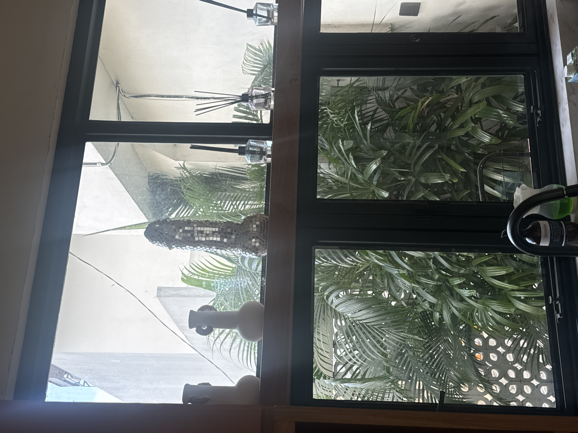 The image shows a view through a multi-paned window in a building located in North Kuta, Indonesia. The window frame is dark and the panes offer contrasting views. The panes on the right side of the frame reveal lush green foliage, including palm trees, which appear to be viewed through a patterned wall, possibly a decorative concrete screen. The panes on the left show a lighter, brighter outdoor setting with a mosaic sculpture, a couple of ceramic vases, and a glimpse of what appears to be the sky and distant sea. There are also details of the building's interior architecture and window fixtures visible. The scene captures a blend of natural and architectural elements, both indoors and outdoors.