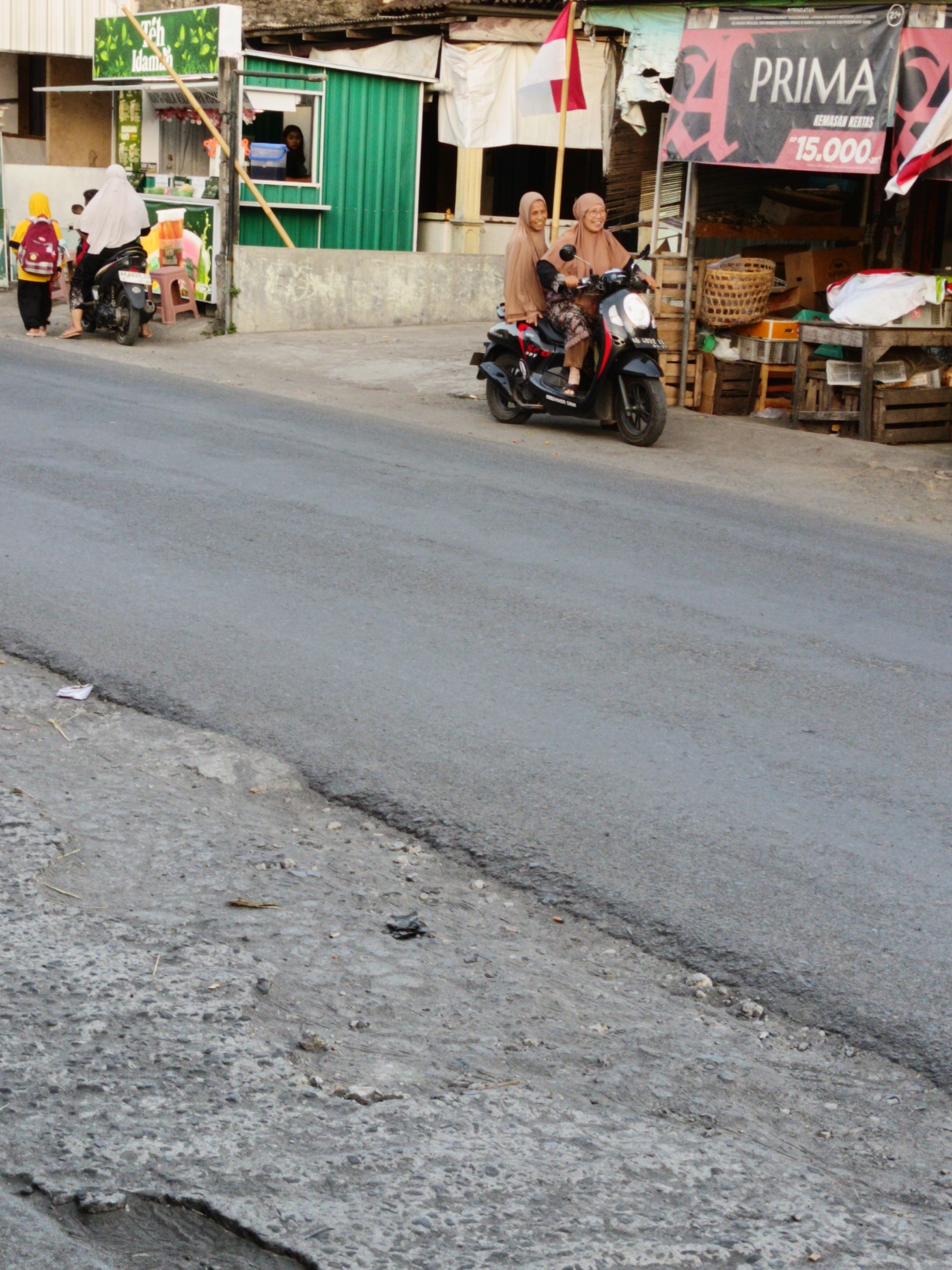 In Sukorini, Indonesia, two women in headscarves ride a black motorcycle on a paved road. The woman in the front is driving, while the woman behind her is smiling. They are both wearing light brown headscarves. The motorcycle is black with red accents. To the right of the motorcycle, there is a small roadside market stall with baskets and wooden crates. A banner with the word "PRIMA" is visible above the stall. To the left of the motorcycle, a person wearing a yellow headscarf and a red backpack stands near a scooter. In the background, a building with green siding and a white awning is visible. There are also several other structures and a flag. The road appears to be in a state of disrepair with cracks and patches.