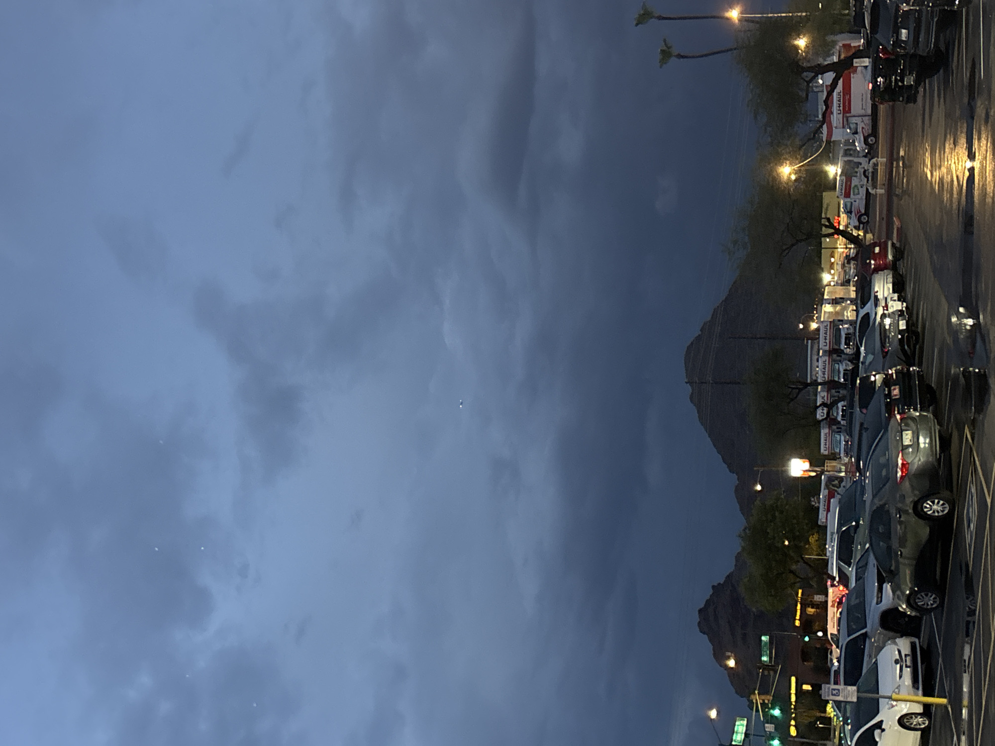 The image captures a twilight scene in Phoenix, Arizona, with a dark, cloudy sky and distant mountains. The foreground features a wet parking lot filled with various cars and U-Haul trucks. Streetlights and building lights illuminate the scene, reflecting off the wet pavement. The overall atmosphere is somber and moody, with the low light suggesting either dawn or dusk. The presence of U-Haul trucks indicates a commercial or residential area, possibly a storage facility or a moving company's hub. The mountainous backdrop is a characteristic feature of the Phoenix landscape.
