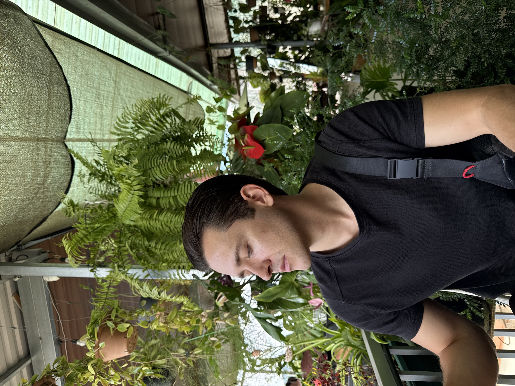 A medium, eye-level shot depicts a man with dark hair wearing a black t-shirt and carrying a black bag with a red detail on its strap. The man appears to be standing among lush greenery in a greenhouse or covered plant area. The location is suggested to be Da Lat, Vietnam, based on the variety of plants and the greenhouse style. Ferns, flowering plants with red blooms, and various other leafy plants create a dense and vibrant environment around the man. A shade cloth or awning is visible above the plants, providing diffused light. The structural elements of the greenhouse, including support beams and plant containers, are also visible. The man's eyes are closed, suggesting a moment of rest or contemplation amidst the foliage.