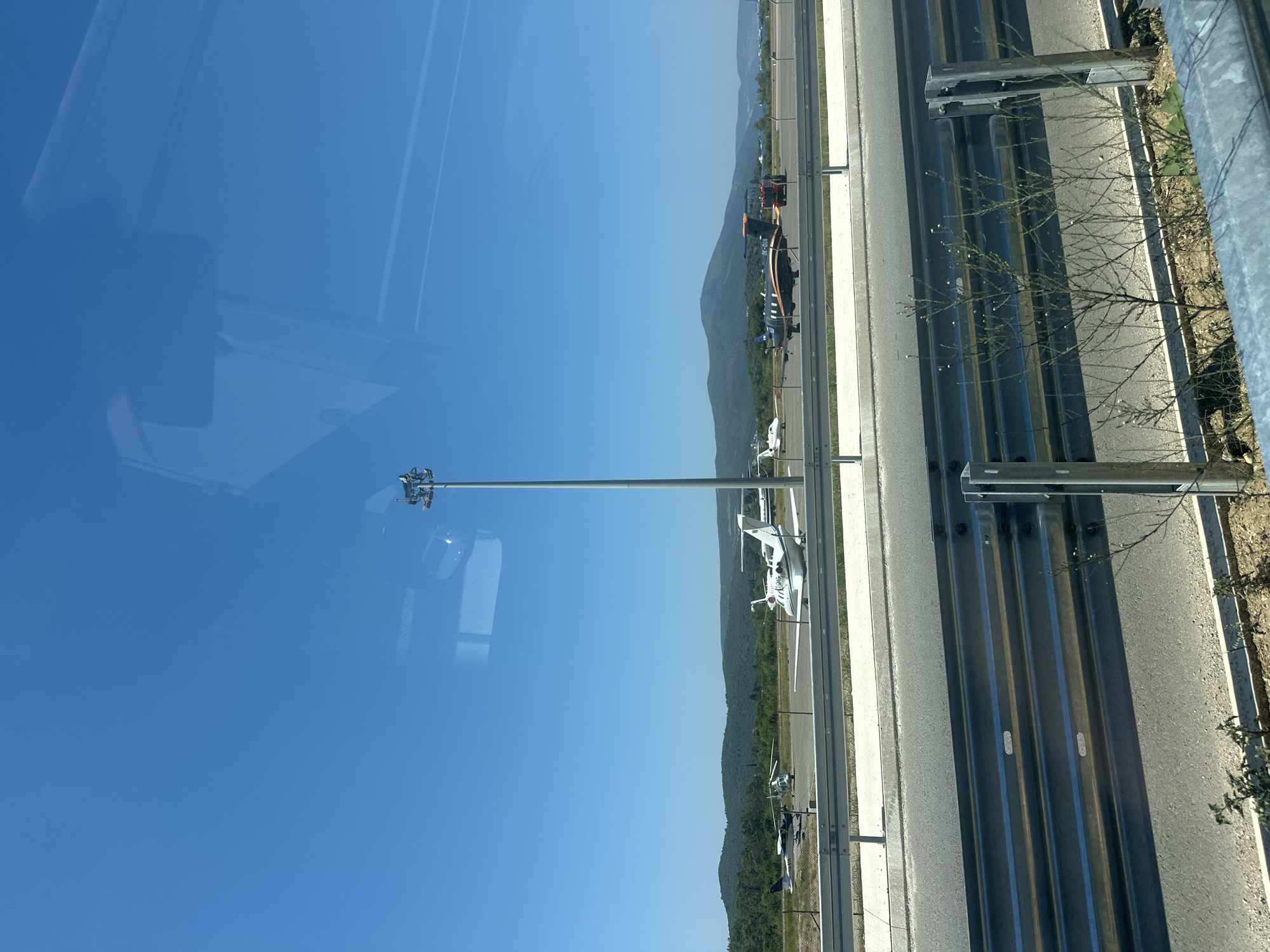 The image shows an airport tarmac under a clear blue sky. Several small aircraft, including a white jet and a dark colored jet with orange accents, are parked on the tarmac. A helipad is visible with a helicopter. In the background, a line of green hills and mountains forms the horizon. A tall lamppost stands prominently on the tarmac, equipped with lights. A reflection of what appears to be a vehicle's interior, possibly a car or bus, is visible on the windshield through which the photograph was taken. A metal guardrail runs across the foreground, separating the roadway from the airport area. The scene suggests an operational small airport. The location is identified as the airport in Mrčevac, Montenegro.