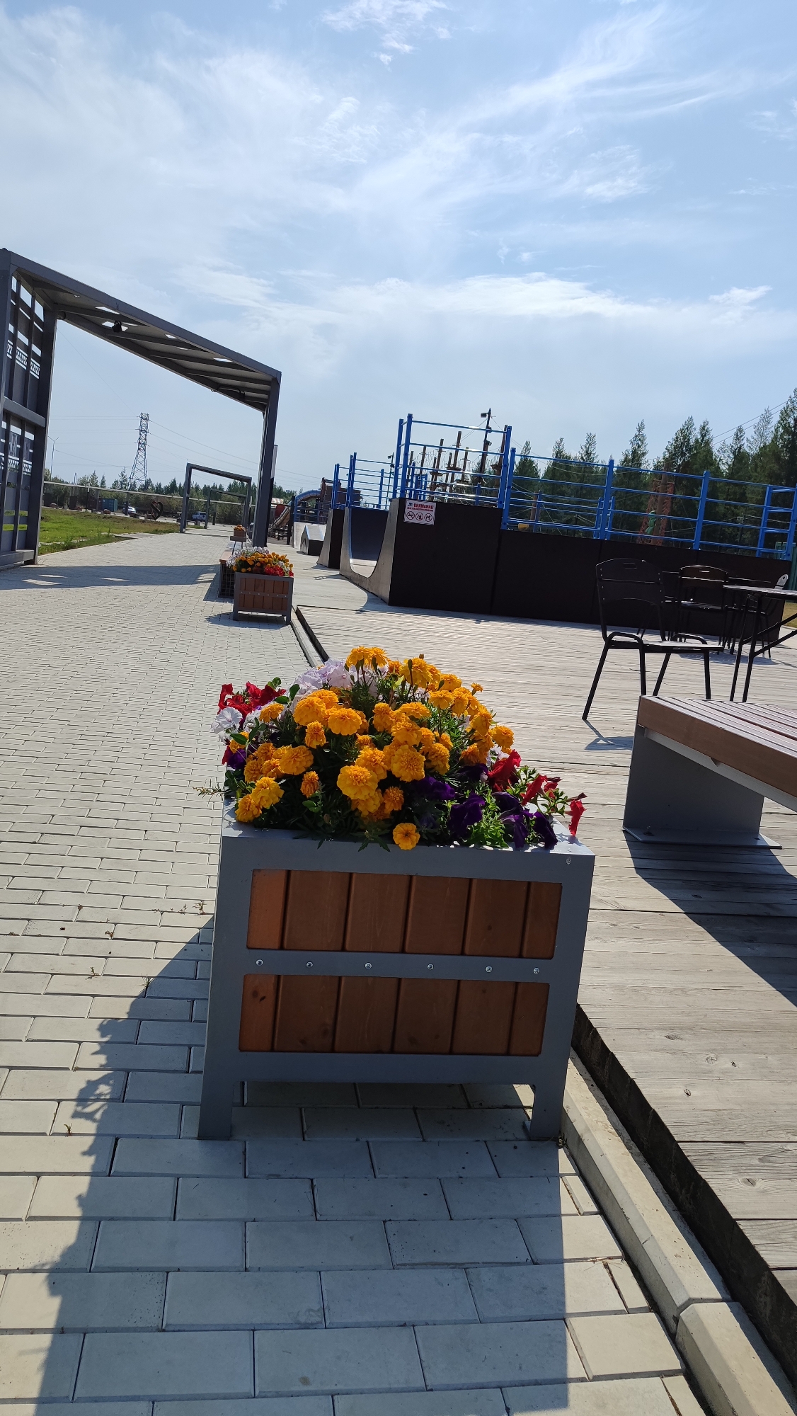 The image depicts an outdoor scene with a focus on a large planter box filled with vibrant yellow and orange flowers, accented by touches of red and purple. The planter, constructed with a grey metal frame and wooden slats, sits on a paved walkway made of light-colored rectangular bricks. To the right of the walkway is a wooden deck area with a bench and a table with chairs. In the background, there are structures resembling a skate park with ramps and blue railings, along with trees and a power line tower against a partly cloudy sky. The location is identified as Mirny, Russia. There are no people visible in the image. The lighting suggests it is daytime with bright sunlight casting shadows.