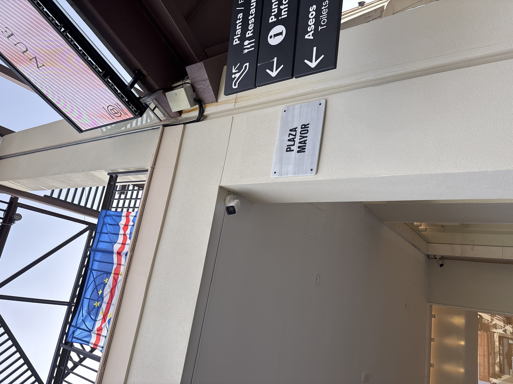 A vertical, eye-level, outdoor, daytime shot captures a section of a building's exterior in Orihuela, Spain. A white, textured wall dominates the left and center of the frame. On the wall, a white marble sign reads "PLAZA MAYOR". To the right of this sign, a black directional sign features white text and icons. The top line indicates "Planta / F" with a fork and knife icon, suggesting a restaurant. The second line reads "Punt" with an information icon. The third line reads "Aseos" with "Toilets" below it, and an arrow pointing left. A small, white security camera is mounted high on the wall, near the corner of the ceiling. Above the wall, a portion of a building's roof structure with dark beams is visible, along with a blue flag bearing a star pattern and red and white stripes, possibly the flag of Cape Verde. To the far right, a large digital billboard displays colorful, striped graphics and text, including a "B" inside a circle logo. The overall scene suggests a public area or commercial establishment.