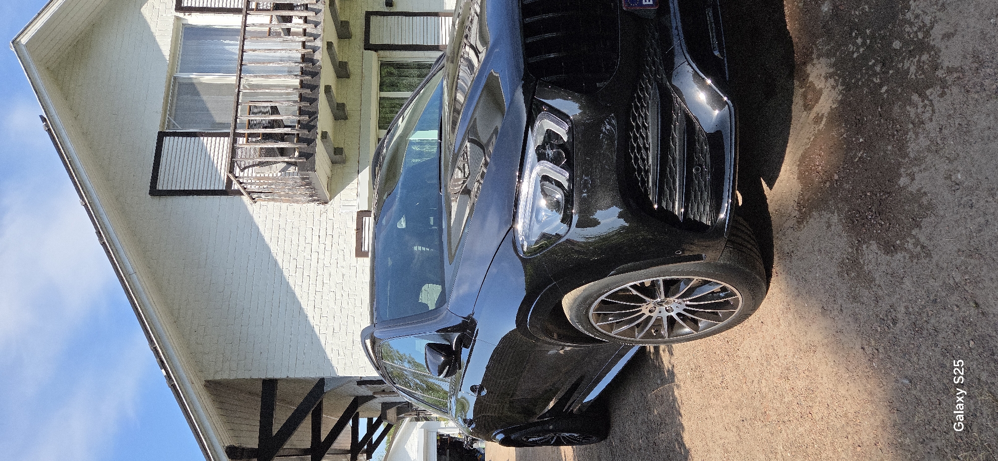 In Eeklo, Belgium, a black Mercedes-Benz GLE AMG SUV is parked on a gravel driveway in front of a two-story white brick house with a balcony. The house has dark shutters on the windows and a dark-paneled roofline. The SUV's front passenger-side wheel and fender are prominently featured, showcasing its intricate alloy rim. The sky above is a clear blue with a few scattered white clouds. The sunlight casts shadows on the ground, indicating it is a bright, sunny day. The overall impression is one of a suburban or semi-rural setting.