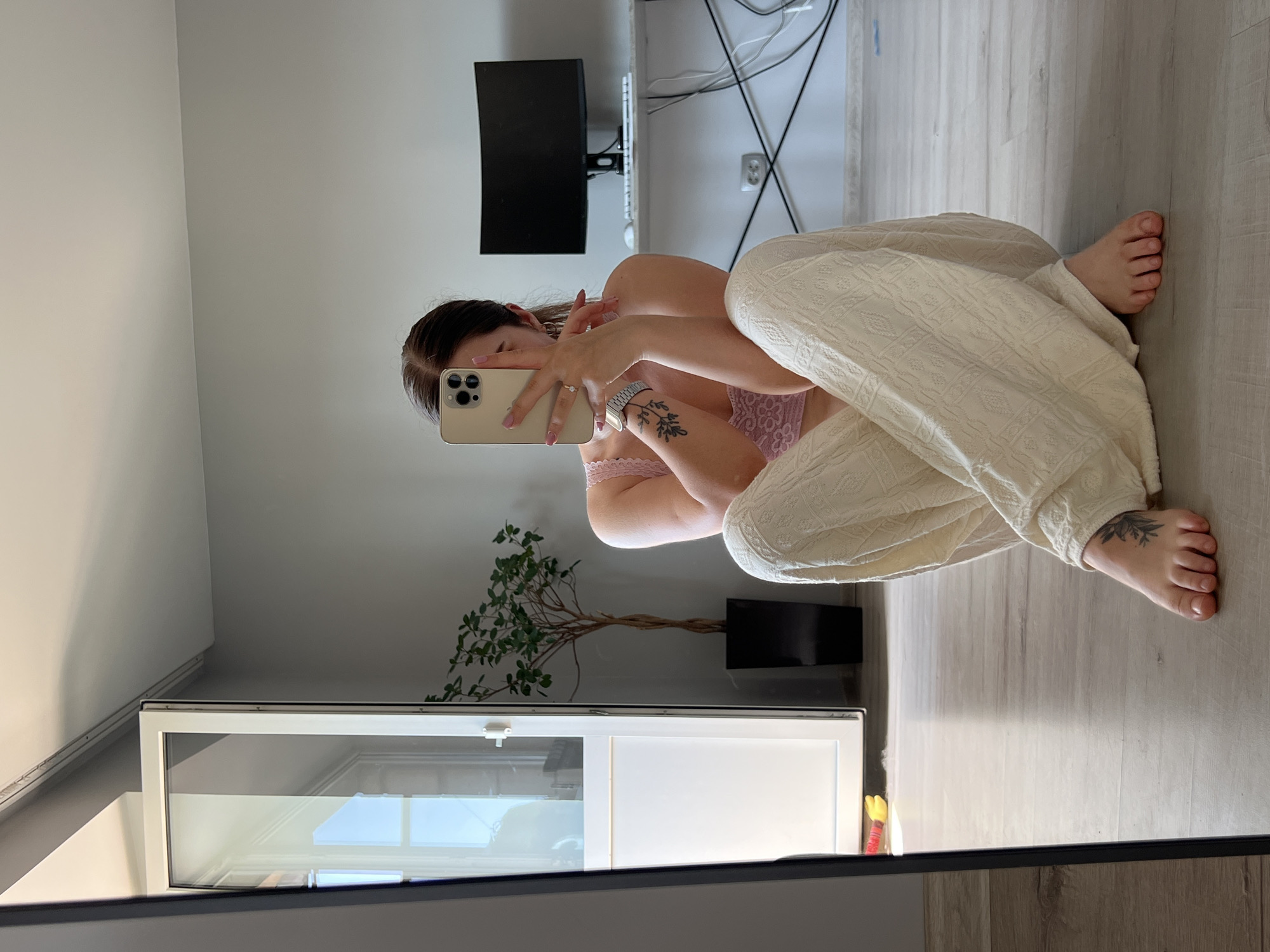 In an indoor setting, a young woman is seated on a wooden floor, taking a selfie with her phone. She is wearing a light pink lace bralette and loose cream-colored patterned pants. Her legs are crossed, and her feet are bare, with a tattoo visible on her left foot. She has jewelry on her wrist and finger.
The room is sparsely furnished. To the left, a tall mirror reflects the scene. In the background, a tall potted plant with green leaves stands in a black planter. A white door with glass panes is partially visible to the left of the plant. Further back, a white desk holds a computer monitor, keyboard, and mouse. Electrical outlets and tangled wires are visible on the wall behind the desk. The walls are painted a light gray, and the ceiling is white. Sunlight streams in from an unseen window, casting shadows and illuminating parts of the floor.
The scene appears to be an apartment or a room in a house. There is no specific indication of the city of Ternopil, Ukraine. The overall impression is of a casual, personal moment captured in a domestic environment.