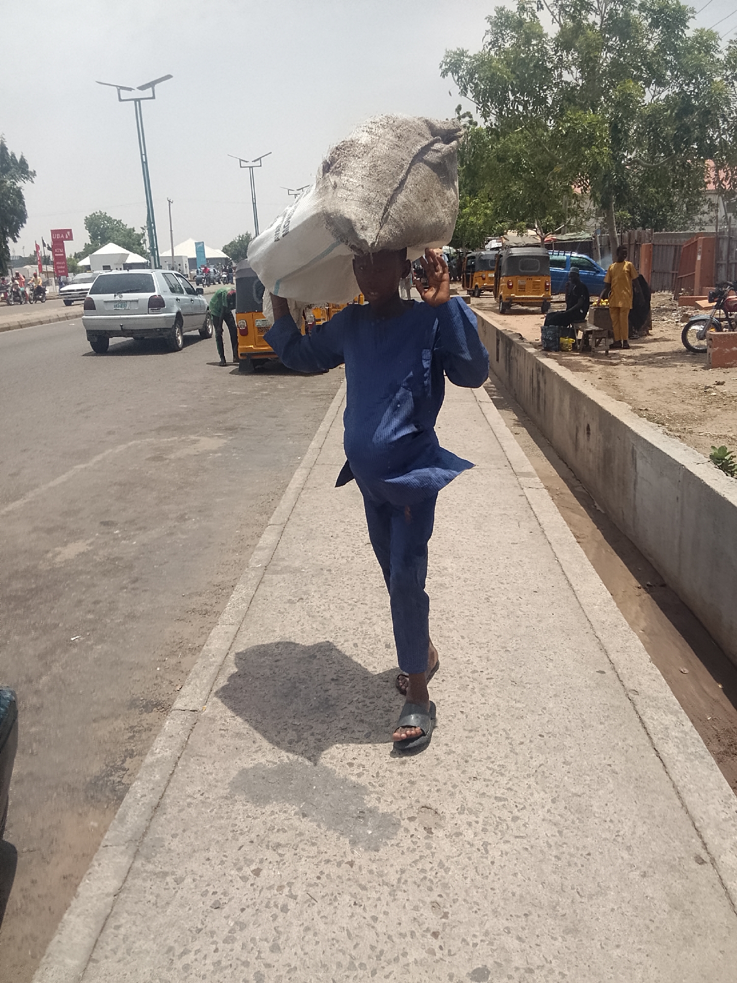 A young man is walking on a concrete sidewalk in a sunny, urban environment in Pantami, Nigeria. He is carrying a large, heavy-looking white sack on his head. The man is wearing a blue, long-sleeved shirt and pants, and flip-flops. His shadow is cast on the sidewalk in front of him.
A busy road is visible to the left of the sidewalk with cars and motorized tricycle taxis. There are pedestrians on the opposite side of the road, and some street vendors. The background includes buildings, trees, and streetlights. A concrete barrier separates the sidewalk from a dirt area. The overall scene is that of a working-class neighborhood.