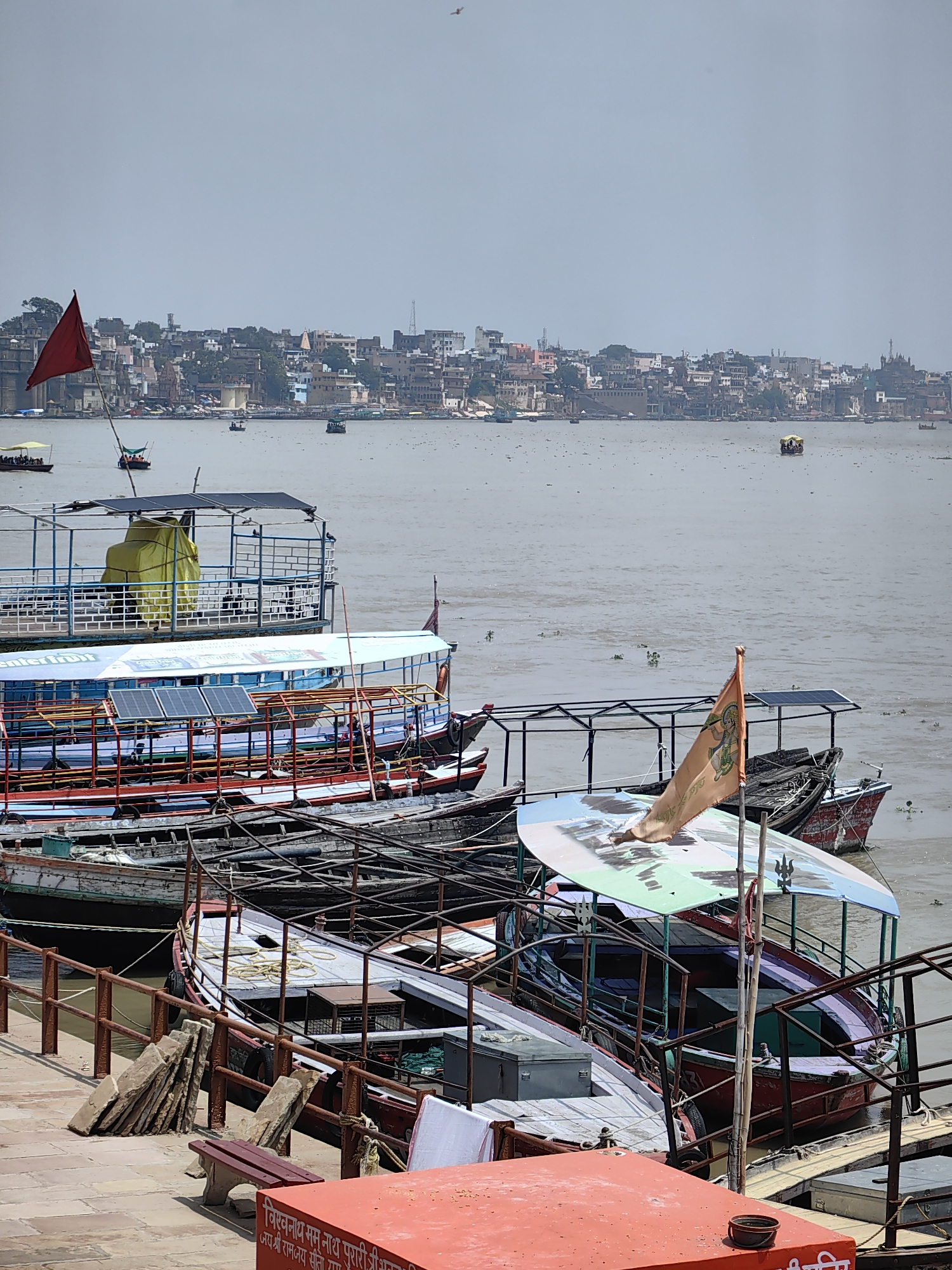This image depicts a scene along the ghats of the River Ganges in Varanasi, India. The foreground shows a collection of moored boats of various sizes and designs. Some boats are covered with awnings, and one prominently displays solar panels on its roof. Several other boats are stacked or moored alongside, with some appearing older and weathered. The water of the Ganges is visible behind the boats, with a distant cityscape lining the opposite bank. The city appears densely built with numerous buildings, temples, and structures, indicative of Varanasi's historical and religious significance. A red flag with symbols is visible on one of the boats. The sky is overcast and hazy, suggesting a warm and humid day. The overall impression is one of a busy waterfront area, likely a hub for river transportation and tourism in this sacred Indian city.