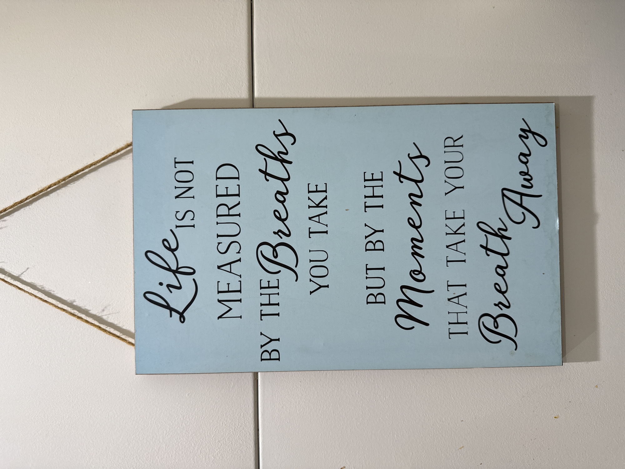 The image shows a rectangular sign with a pale blue background and black lettering, displaying the quote: "Life is not measured by the breaths you take but by the moments that take your breath away." The sign is hung by a thin, beige string attached to the top left corner. The sign is mounted on a wall. The location and context are not possible to ascertain from the image.