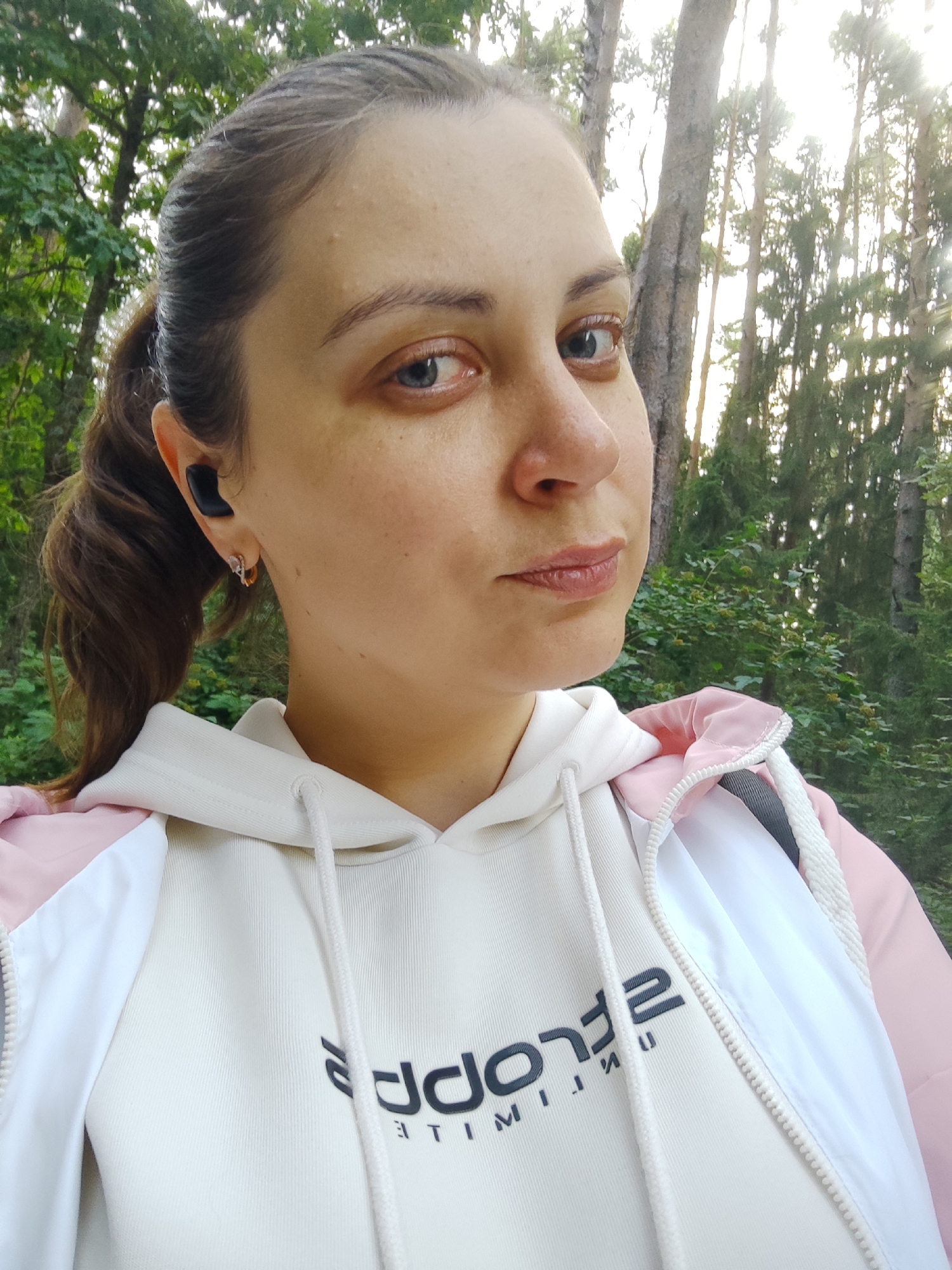 A medium shot captures a woman with her hair pulled back in a ponytail, wearing wireless earbuds and a hoodie with a pink and white jacket over it. She is looking towards the camera with a neutral expression. The background features a forest with tall trees and green foliage, illuminated by sunlight filtering through the leaves.