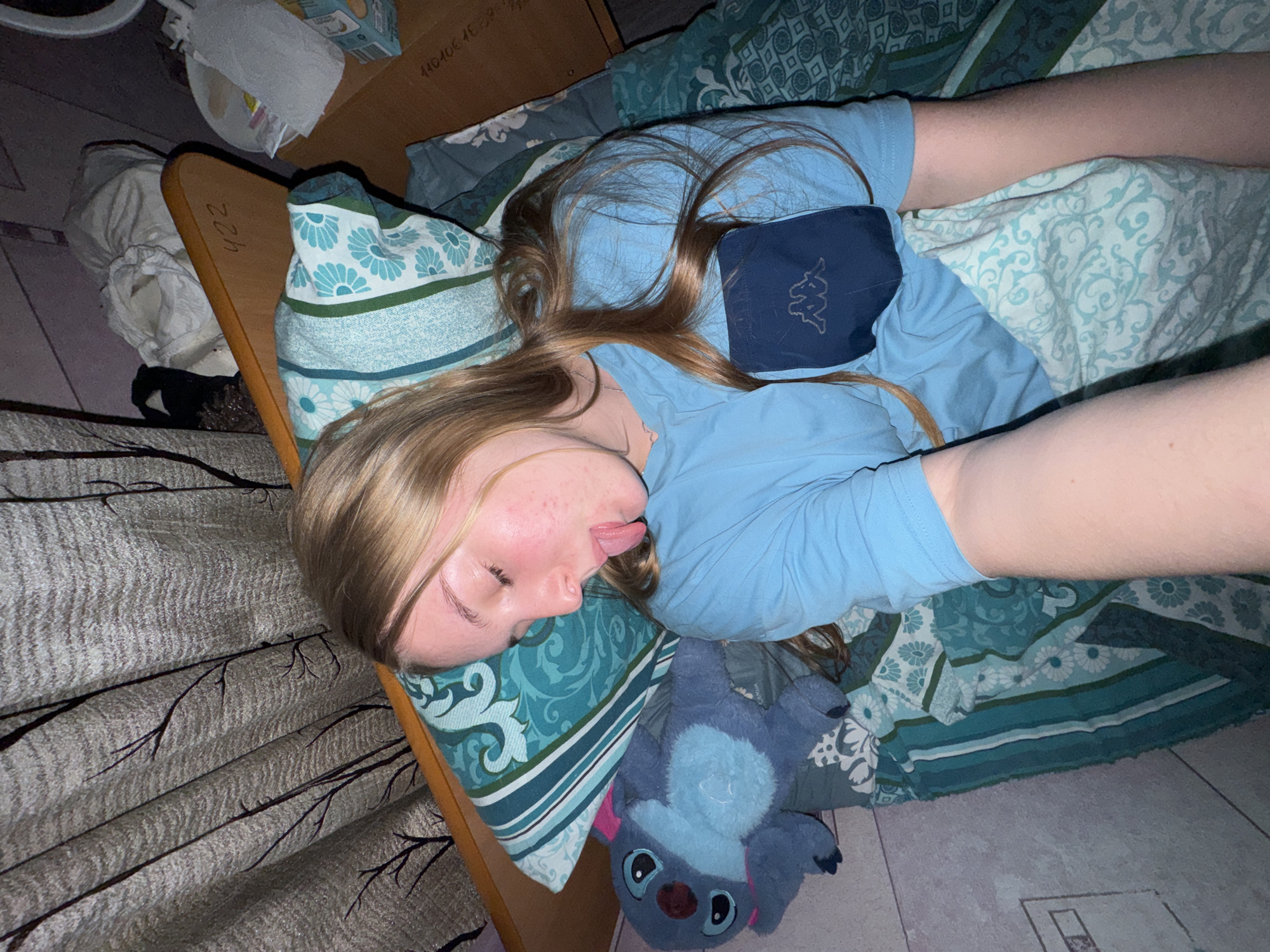 A fair-skinned person with light brown hair is lying on their back in a bed covered in patterned bedding. The person is wearing a light blue shirt and sticking their tongue out. A blue stuffed animal lies next to the person on the bed. The bed is positioned near a wall with dark patterned curtains. The room also contains some clutter, including cardboard boxes and other objects. The image was taken in Perm, Russia.