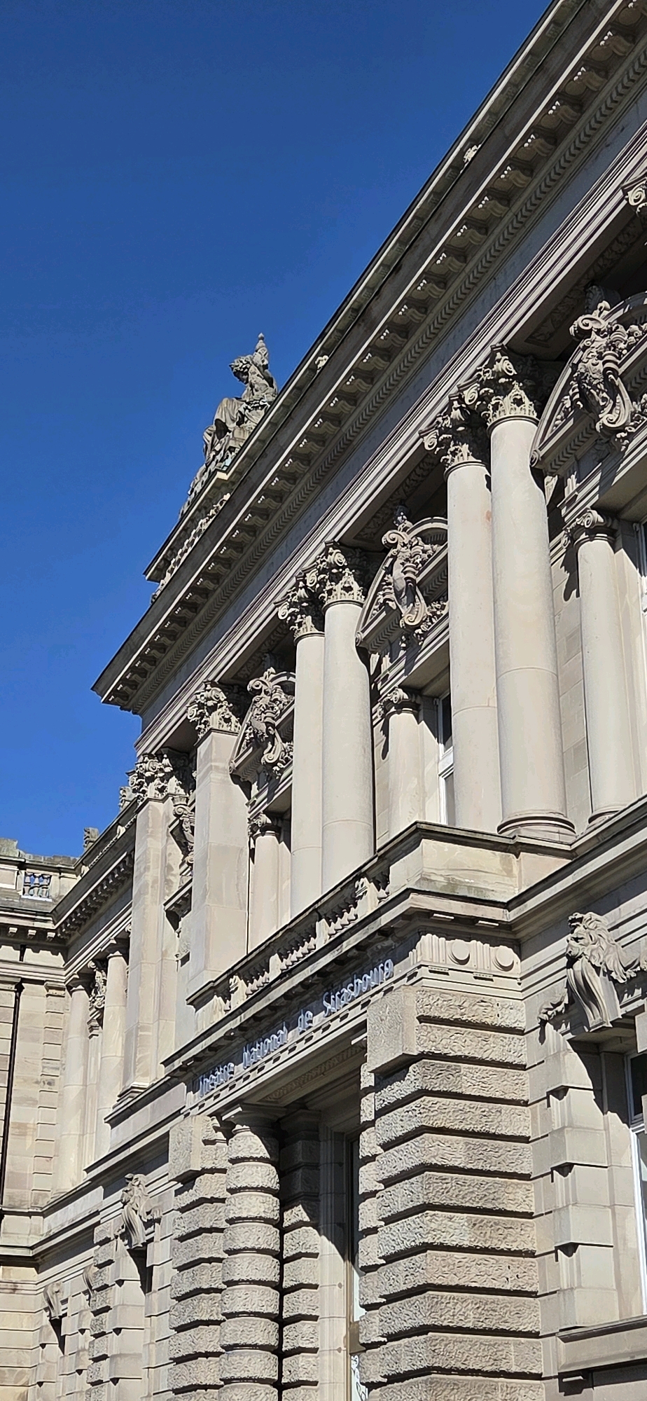 This is a low-angle, upward shot of the exterior of the Opéra National du Rhin in Strasbourg, France. The building is constructed of light-colored stone and features classical architectural elements such as large columns with ornate capitals, decorative friezes, and statues adorning the roofline. The facade is intricately detailed with carvings and moldings. The sky above is a clear, vibrant blue. The sunlight casts strong shadows, highlighting the building's three-dimensional features. The text "Opéra National de Strasbourg" is visible above an entrance.