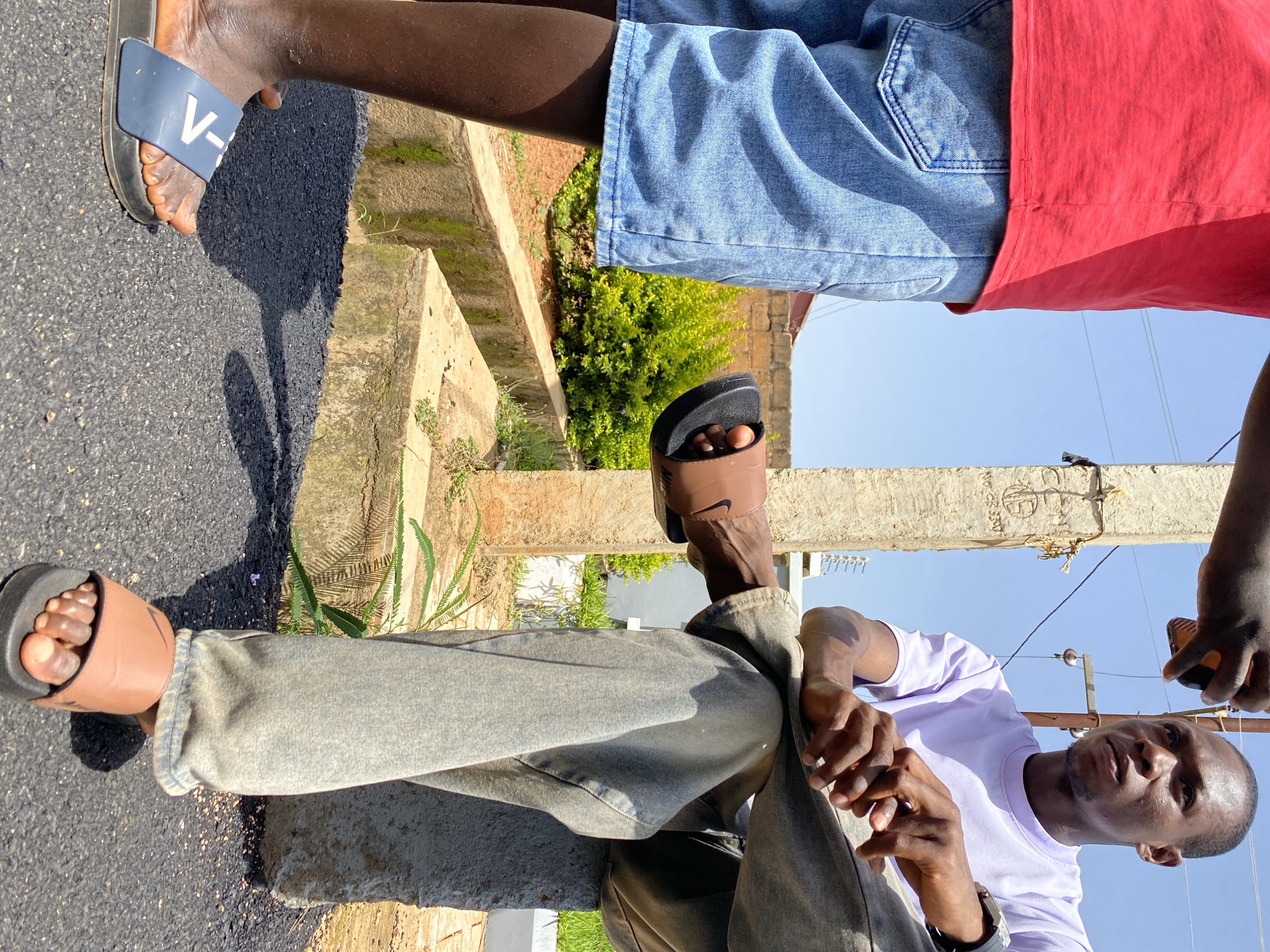 The image shows two Black men in Bukuru, Nigeria. One man, on the left, wears a red t-shirt and denim shorts. The other man, on the right, is seated on a concrete block, wearing a purple t-shirt and grey jeans. He is looking to his right, with his hands resting on his lap. A concrete pillar stands behind him, with utility wires running overhead. The ground is paved with asphalt. In the background, there is a low brick wall and green foliage. The man on the left is wearing blue slides with a white "V-" marking, and the man on the right is wearing brown slides with a Nike logo. The scene appears to be an outdoor setting on a sunny day.