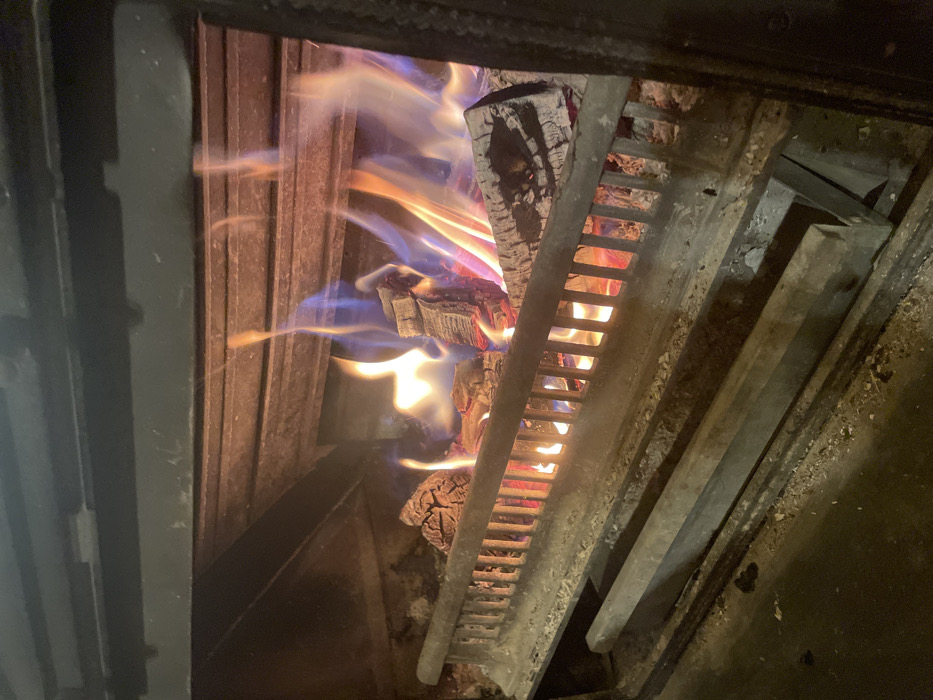 Within an enclosed fireplace, located in Palež, Montenegro, multiple wooden logs are actively combusting. The fire displays bright yellow and orange flames with distinct blue sections, emitting light that illuminates the immediate interior of the firebox. The logs rest on a horizontal metal grate, which features vertical bars in the foreground. Below the grate, an ash pan or drawer mechanism is visible, containing a minimal amount of light-colored ash and dark debris. The interior surfaces of the fireplace are dark, likely metallic or stone, showing signs of soot and heat exposure. No individuals are present in the scene.