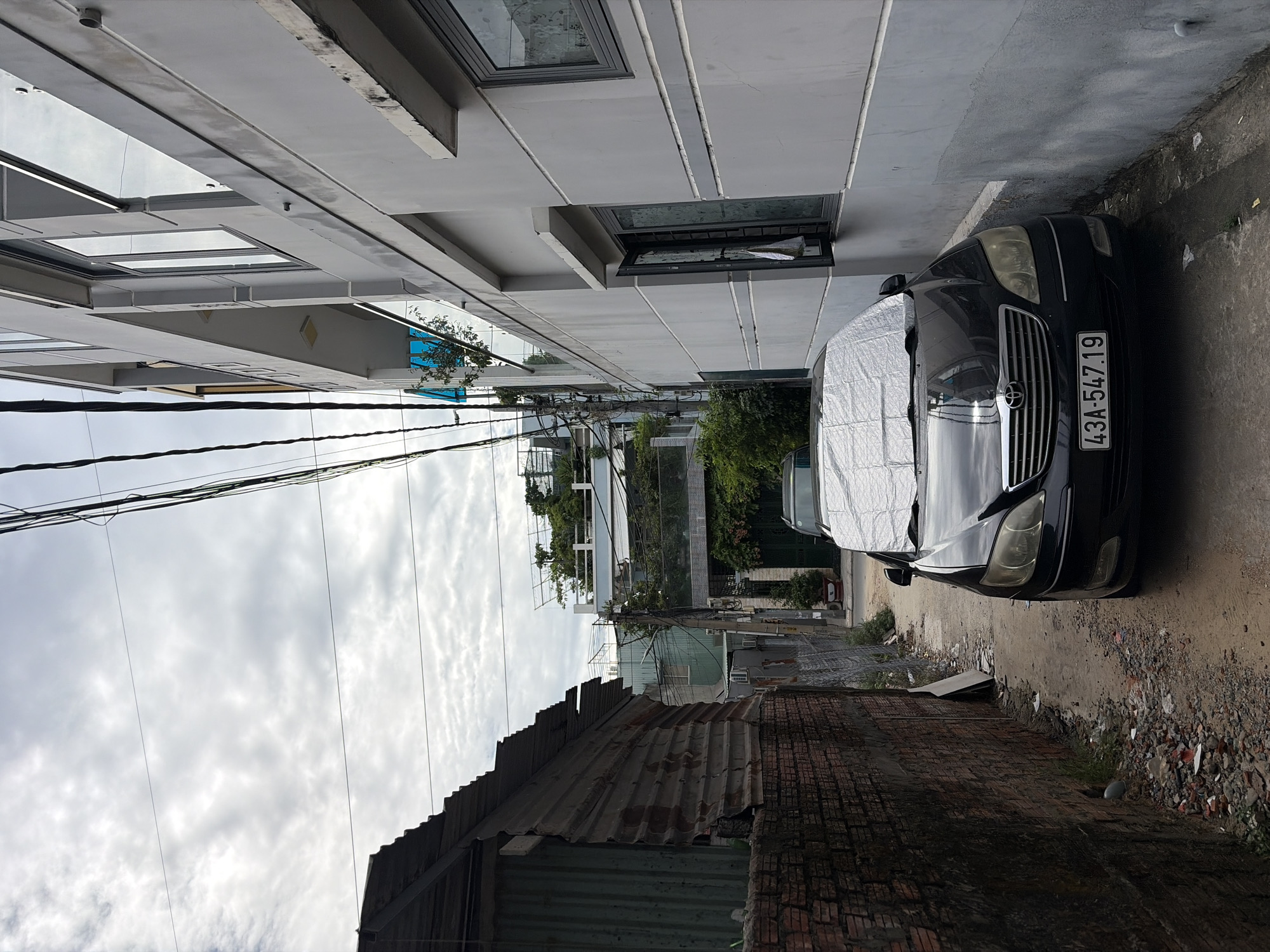 The image shows a narrow alleyway in Đà Nẵng, Vietnam, bordered by a corrugated metal structure and a modern, multi-story building. A black sedan, covered with a reflective car cover on its hood, is parked in the alley. The building on the right has several floors with balconies and windows, some of which are open. Overhead, a tangle of electrical wires crisscrosses the sky, which is overcast with clouds. The ground of the alleyway is dirt and gravel, with some debris scattered around. The corrugated metal structure on the left appears weathered and partially damaged, with a brick wall visible at its base. In the background, more buildings and lush greenery can be seen.