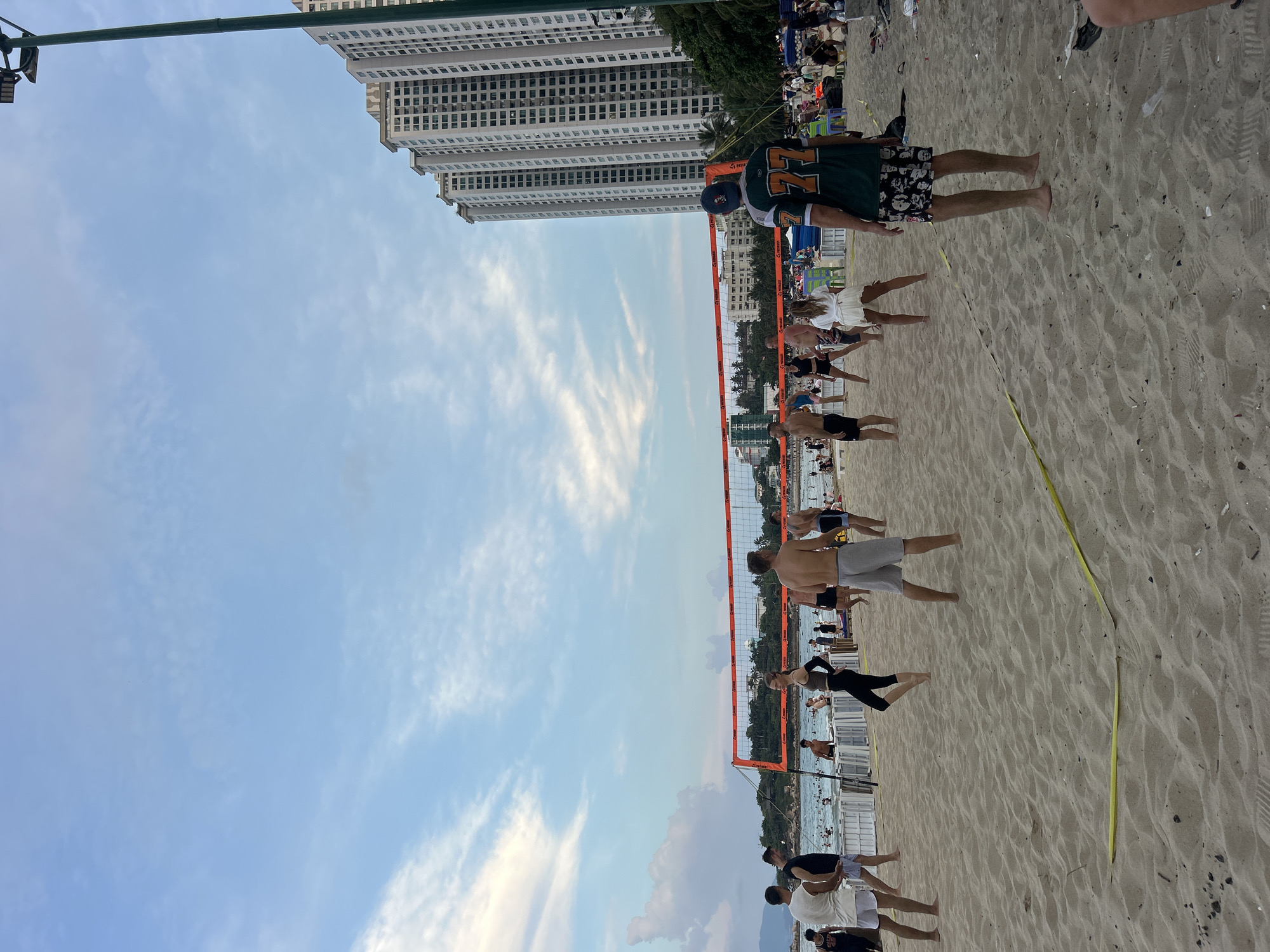 Captured in Bac Nha Trang Ward, Vietnam, the image presents a beach volleyball game in progress. Several individuals stand or move on the sand court, delineated by an orange net and a yellow boundary line. In the background, apartment complexes rise, complementing the sky filled with streaky clouds. The beach and ocean are visible with people in the water. In the upper right corner of the photo, several people are sitting in chairs.