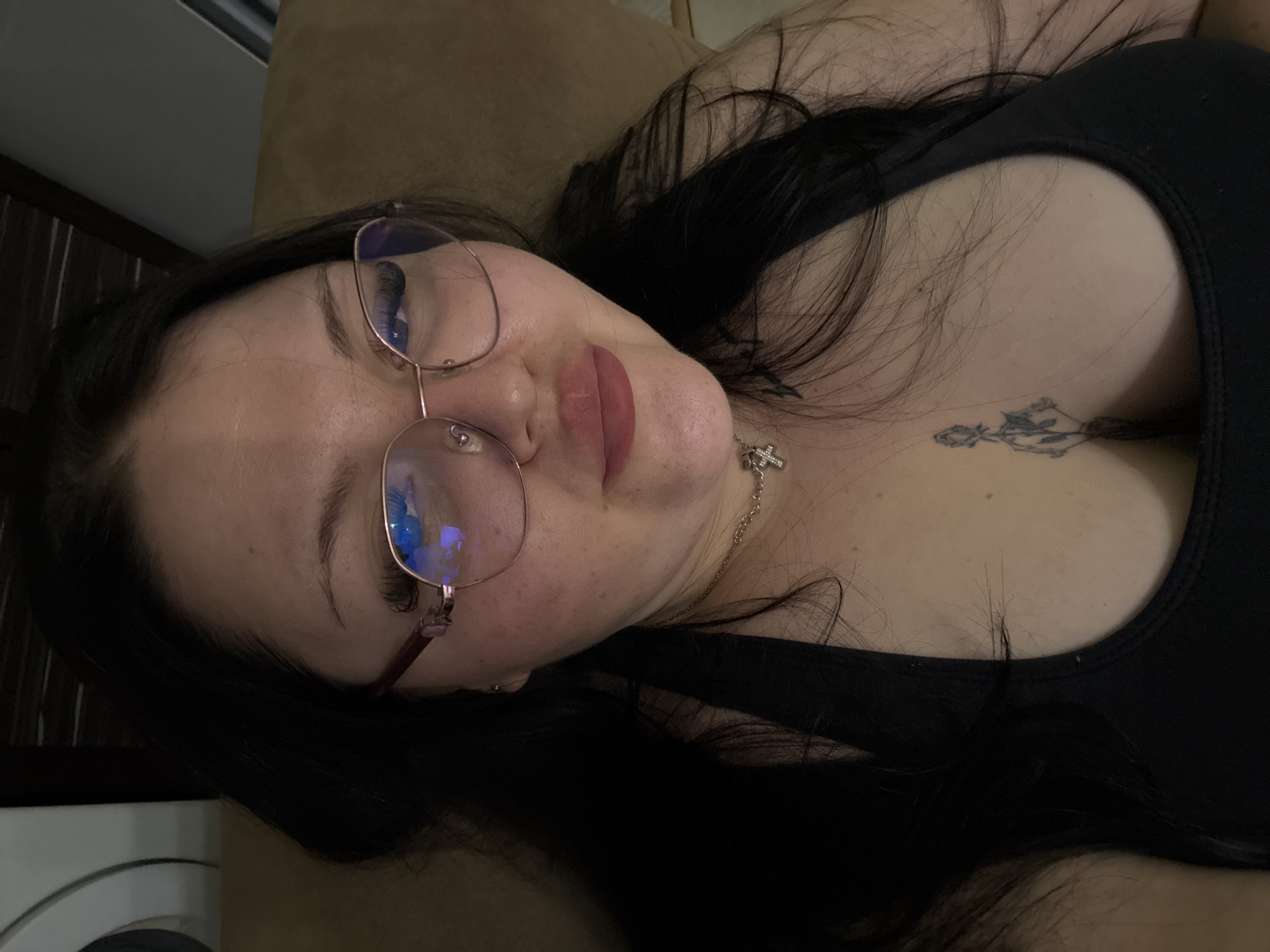 A person with long, dark hair is reclining, wearing glasses and a black tank top. They have a tattoo on their chest and are wearing a necklace with a cross pendant. The background appears to be a couch or bed. There is no indication of a location in Sofia, Bulgaria.