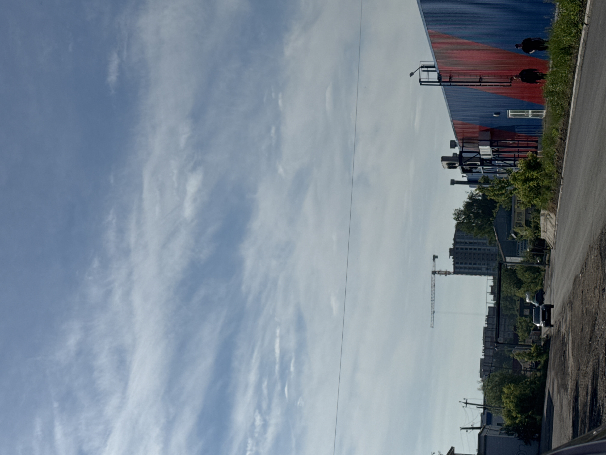 The image depicts an urban or industrial landscape under a bright, sunny sky with wispy white clouds. The perspective is slightly elevated, looking down a road or alleyway.
On the left side, a large building dominates, featuring a corrugated metal exterior painted in two distinct sections: red on the upper part and blue on the lower. A white rectangular window is visible on the blue section. Attached to the building's facade is a dark metal structure, possibly a set of stairs or a utility platform, topped with a street lamp. Two dark, indistinct figures, possibly people or objects, are visible near the top edge of the red section of the building, appearing to be on the roof or an upper level. Green vegetation, including grass and bushes, lines the base of this building and the adjacent road.
To the right, a road, possibly unpaved or heavily worn, curves into the foreground. In the midground and background, an urban environment unfolds. A prominent yellow construction crane stands tall, indicative of ongoing development. Behind the crane, a large, multi-story building, possibly residential or commercial, is visible. Further down the road, several cars are parked or moving, and power lines strung between poles crisscross the scene. Various other buildings and structures, along with scattered trees and bushes, populate the background.
The time of day appears to be daytime due to the bright illumination and clear sky. A distant sign in the background bears partially legible Cyrillic text, which seems to say "КУПИМ ВТОРИЧНОЕ СЫРЬЕ" (BUY SECONDARY RAW MATERIALS), suggesting a recycling or industrial business in the vicinity. There is no overt activity or event taking place, presenting a static snapshot of the location.