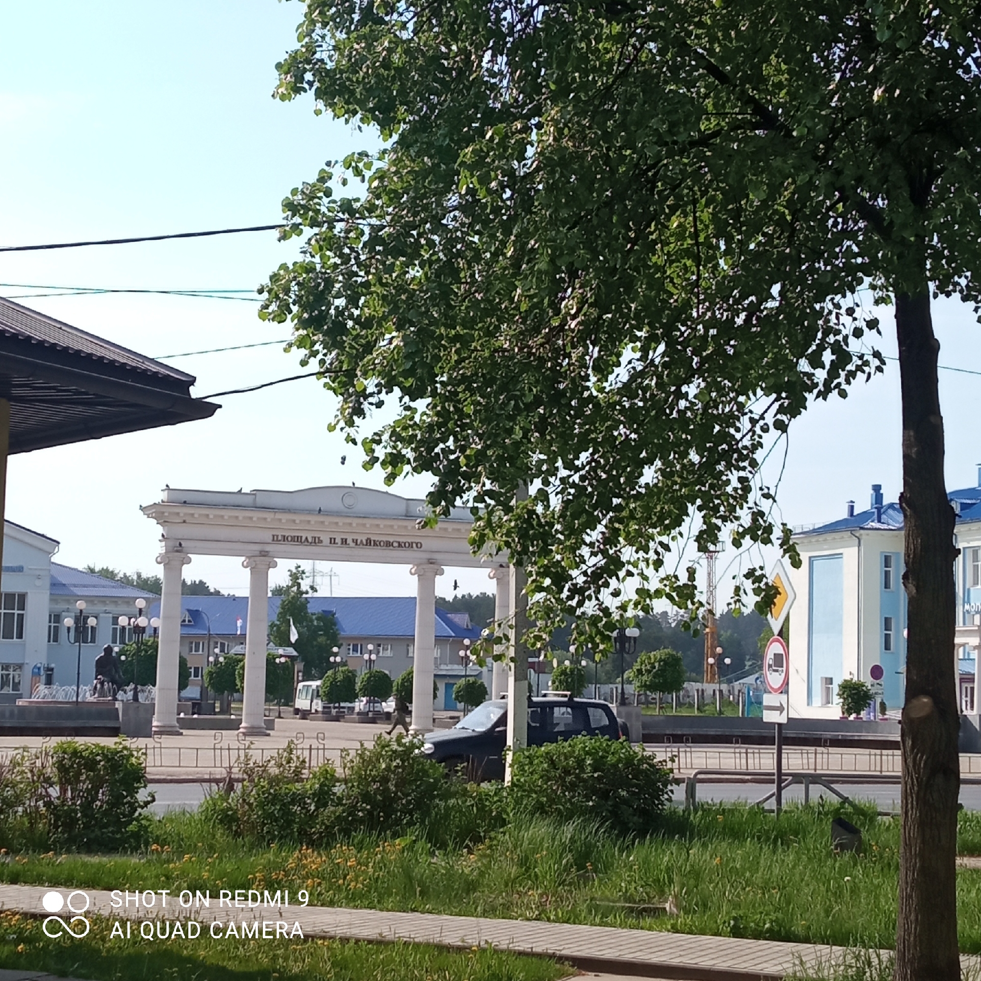 The image shows a street scene in Chaikovsky, Russia, featuring the "Ploshchad P.I. Tchaikovskogo" (Tchaikovsky Square). A white archway with columns, bearing the inscription "ПЛОЩАДЬ П. И. ЧАЙКОВСКОГО" (P.I. Tchaikovsky Square), is visible in the background. There are buildings with blue roofs and white walls, as well as street lamps and trees in the square. A dark-colored car is parked on the street. A road sign indicating "No Trucks Allowed" is positioned on the right side of the frame. A mature tree with green foliage dominates the right side of the image. In the foreground, there are patches of green grass and plants. A paved walkway is visible at the bottom of the image. Power lines stretch across the sky. The image is taken during daylight hours under a clear sky.