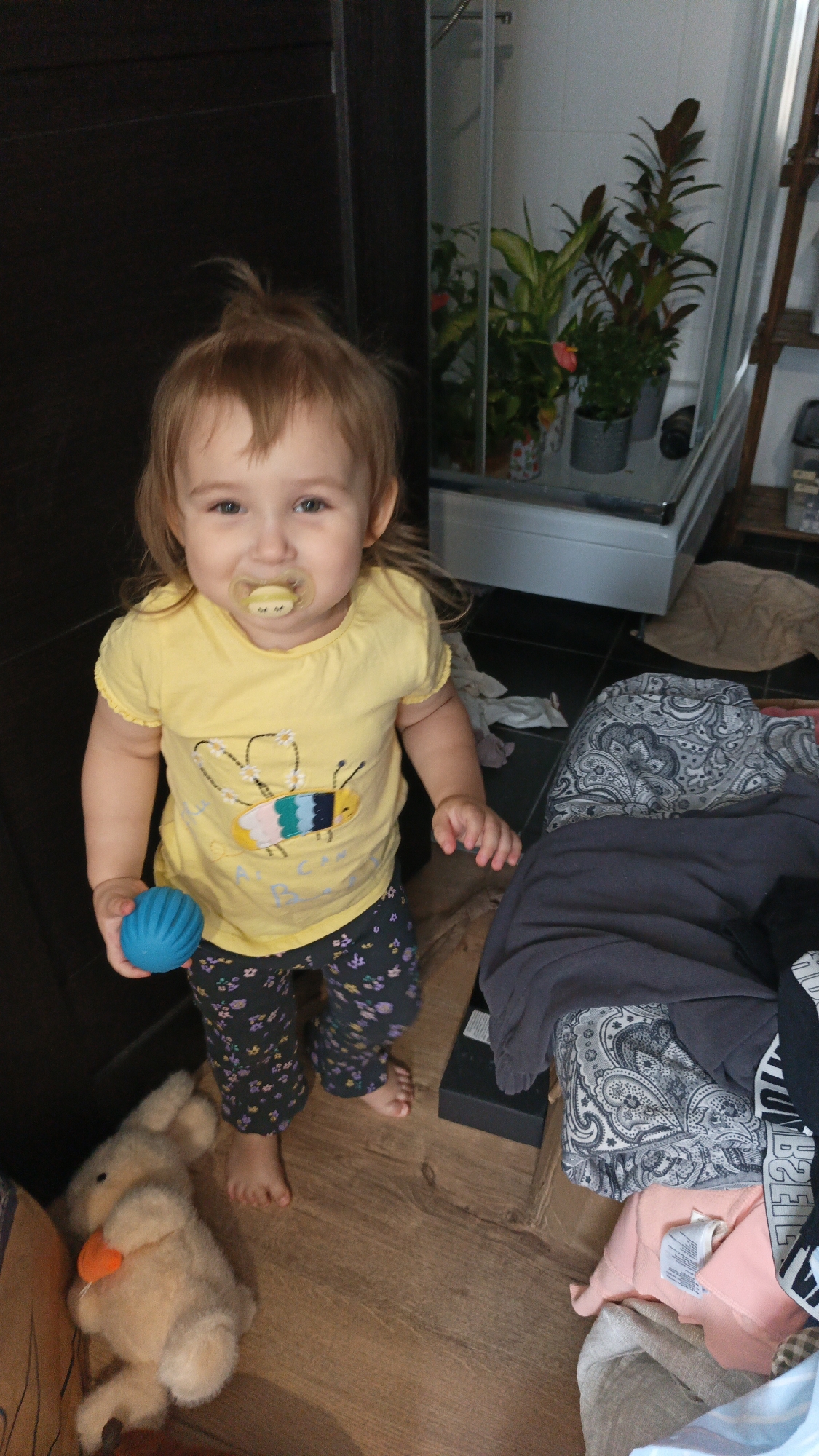 A young Caucasian girl, estimated to be between 18-30 months old, stands barefoot on a wooden floor indoors. She has light brown hair styled loosely and a pacifier in her mouth. She is wearing a yellow t-shirt with a bee graphic and dark pants with a floral pattern. In her left hand, she holds a small, blue, textured ball. To her right, on the floor, is a light brown stuffed rabbit with an orange carrot. The background shows a shower enclosure with several potted plants, and to the right, a shelf unit with items. The floor is cluttered with various pieces of clothing, including a dark grey garment and a patterned textile. The overall environment appears to be a home setting. The image was taken on August 21, 2021, at 3:25 PM in Balashikha, Russia.