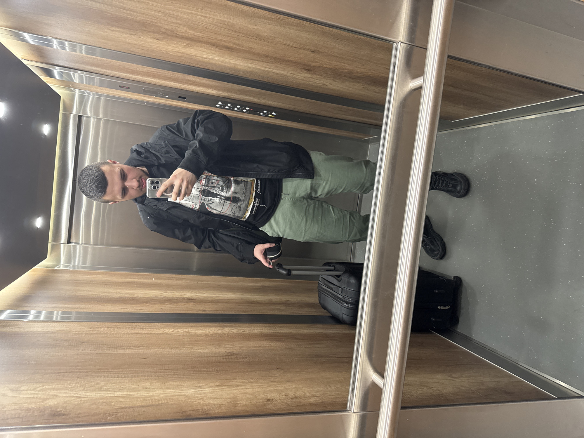 A man takes a mirror selfie inside an elevator. He is positioned slightly off-center to the left, capturing his reflection. He wears a black jacket over a black t-shirt featuring a white graphic print that includes what appears to be a map or architectural drawing and partially visible, reversed text such as "THE LIVING...". His lower half is clad in green or khaki pants and black shoes. He holds a silver smartphone, likely an iPhone, in his right hand to take the picture, and a black cup or thermos in his left. His gaze is directed towards the phone screen, displaying his reflected face.
The elevator interior features warm-toned wooden panels on the walls, segmented by horizontal metallic strips. A panel of illuminated elevator buttons is visible on the wall behind the man's head, though individual button details are indistinct. Polished metallic handrails run along the mirrored surface. The floor is a dark gray with light speckles. Next to the man's reflection, on the right side of the image, a small black rolling suitcase stands on the floor, indicating travel. The scene conveys a moment of transit in a modern, well-maintained indoor environment. The context provided is Sofia, Bulgaria.