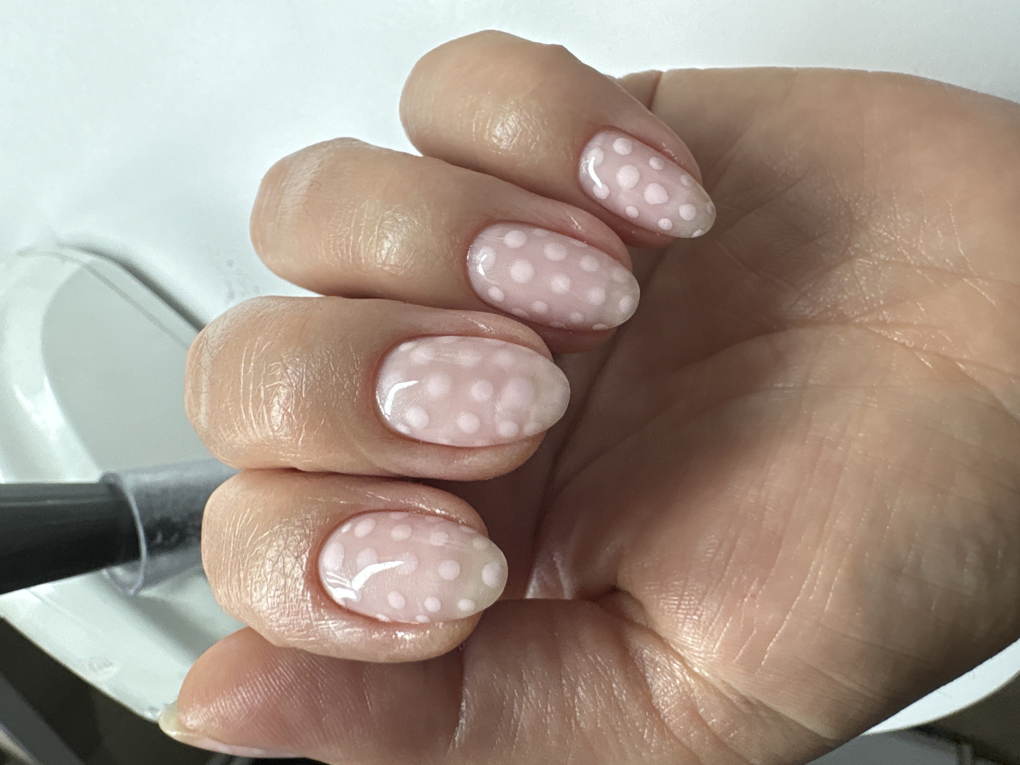 The image depicts a person's hand with manicured nails painted in a light pink color with white polka dots. The fingers are slightly curled, with the nails prominently displayed. Part of a clear container with a black lid is visible in the lower left corner. The background appears to be a plain, light-colored surface. There is no information to indicate the location is Kyiv, Ukraine.