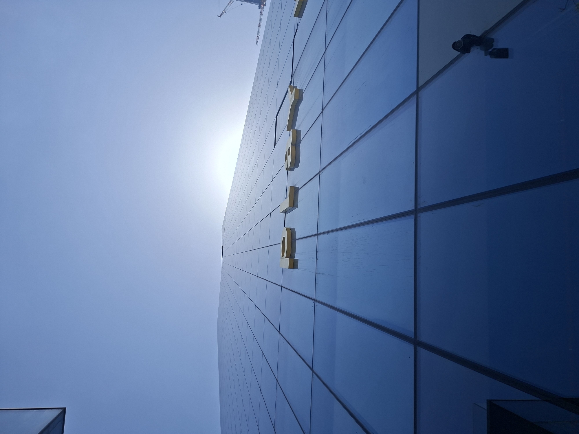From a low-angle perspective, the image captures a modern, high-rise building in Tel-Aviv, Israel. The building is predominantly blue and features a grid-like design with numerous rectangular glass panels. A vertical line of large, gold-colored letters or symbols is affixed to the building’s facade, starting from the upper portion and extending downwards. Near the top-right corner of the building, a black security camera is visible. In the background, against a light blue sky, a construction crane is partly visible, suggesting ongoing development in the vicinity. The sun is also visible, creating a bright glare in the sky.