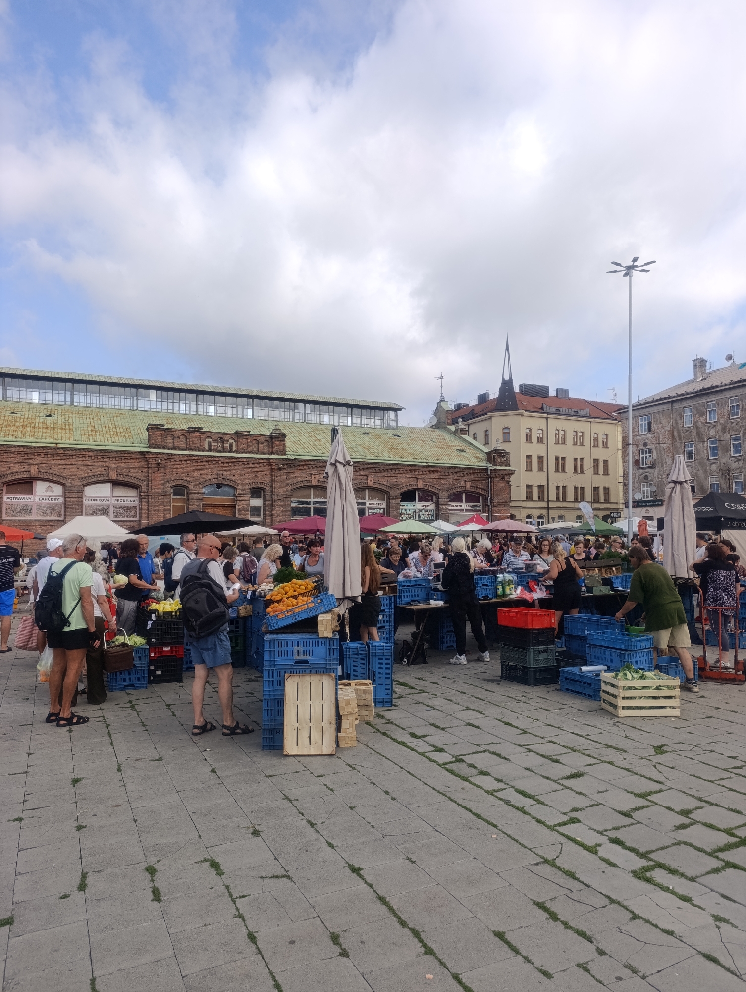 The image shows a busy outdoor market scene in Olomouc, Czechia. A variety of people are browsing stalls and purchasing goods. Stalls are laden with fresh produce, including crates of oranges and other fruits and vegetables. Several blue plastic crates are stacked, containing various items. Umbrellas provide shade over some of the market areas. In the background, there are large brick buildings with arched windows, characteristic of older European architecture, and a taller building with a distinctive spire. The ground is paved with square concrete slabs, with some patches of grass growing in the cracks. A tall lamppost with multiple lights is visible in the sky. The sky is partly cloudy.