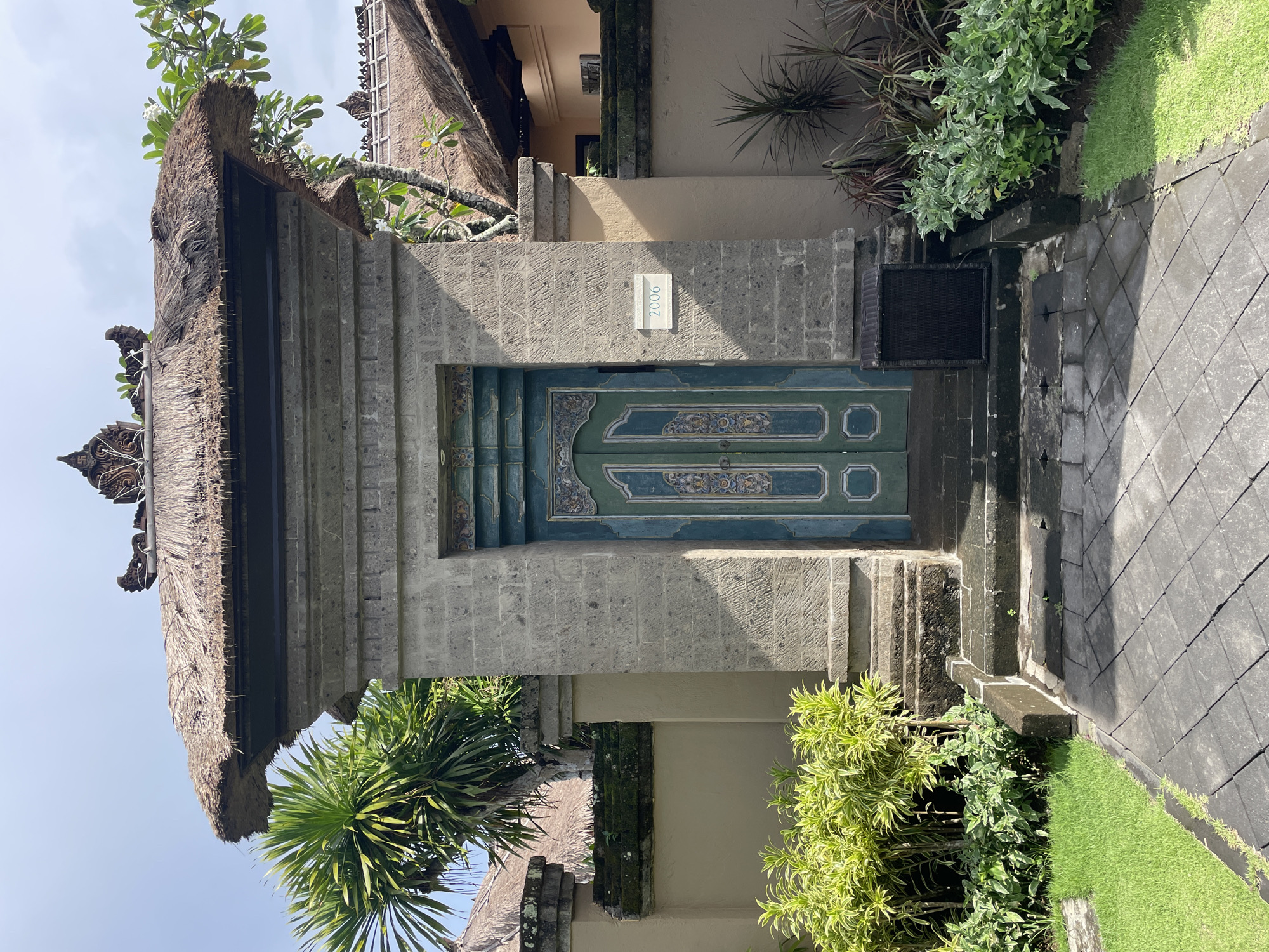 The image depicts a traditional Balinese entrance gate in Kuta Selatan, Indonesia. The gate features a thatched roof and a stone structure framing a wooden double door painted in turquoise and decorated with ornate carvings. The address "2006" is visible on a sign near the top of the gate. Vegetation, including trees and shrubs, surrounds the gate, and a paved path leads to the entrance.