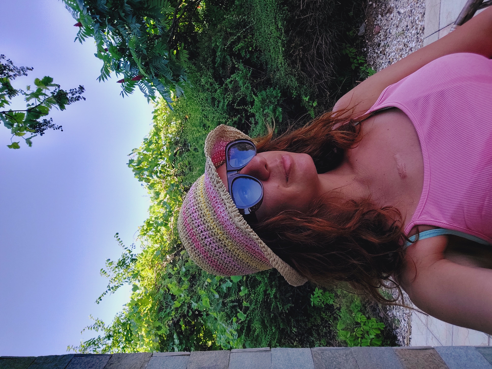 A woman wearing a pink tank top and a straw hat with pink and yellow stripes, accessorized with large sunglasses, is captured from the chest up. She has wavy, reddish-brown hair and appears to be looking to her right. The background consists of lush green foliage under a clear blue sky. A part of a stone structure is visible on the left side of the frame. The setting suggests an outdoor location with abundant greenery.