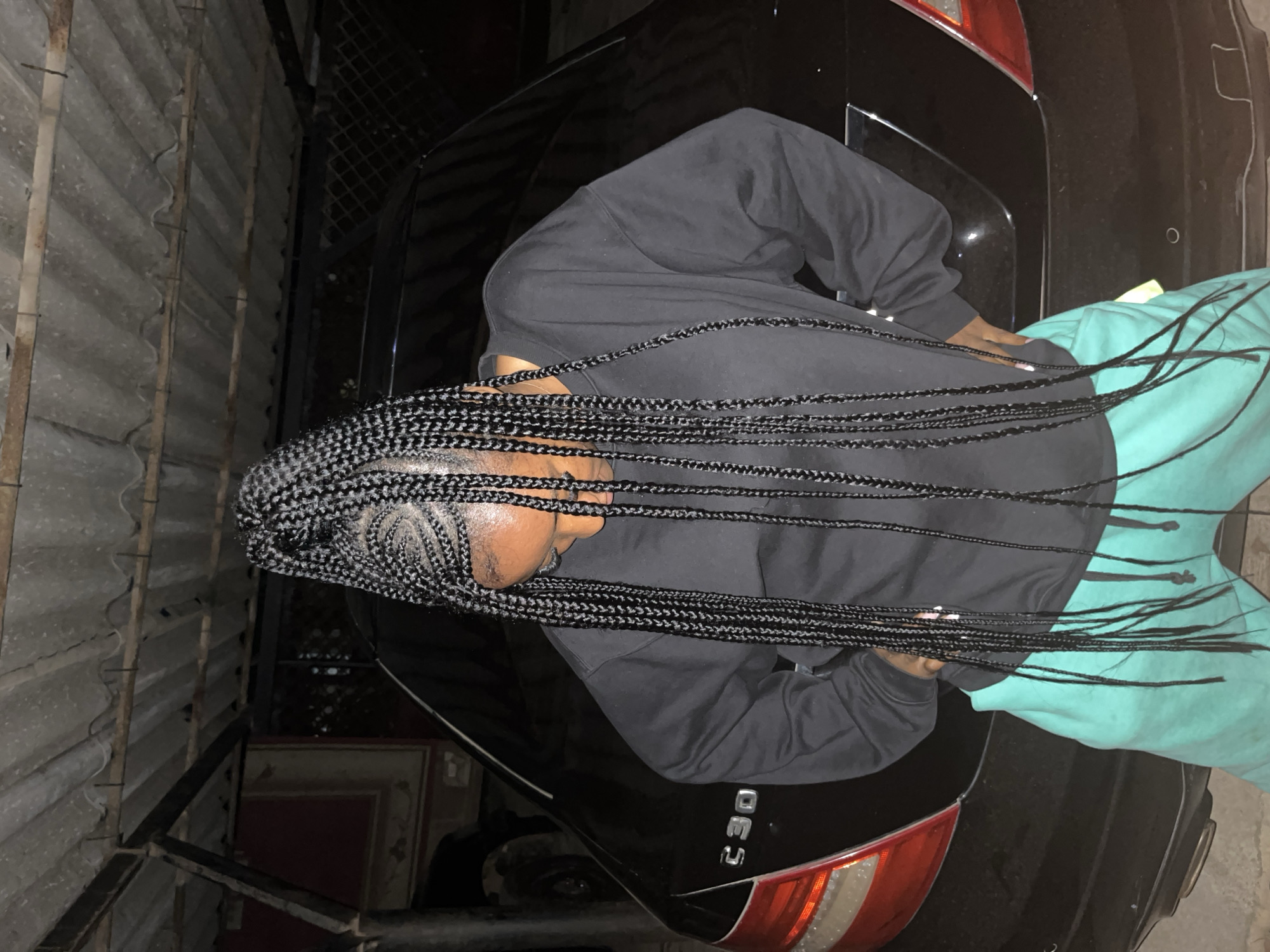 A person with long, braided hair stands in front of a black Mercedes-Benz C300 car. The person is wearing a black long-sleeved shirt and teal sweatpants. The background shows a corrugated metal roof supported by metal beams. The scene appears to be an outdoor parking area, possibly in Jos, Nigeria, given the reference to the city. The lighting suggests it is nighttime or dusk.