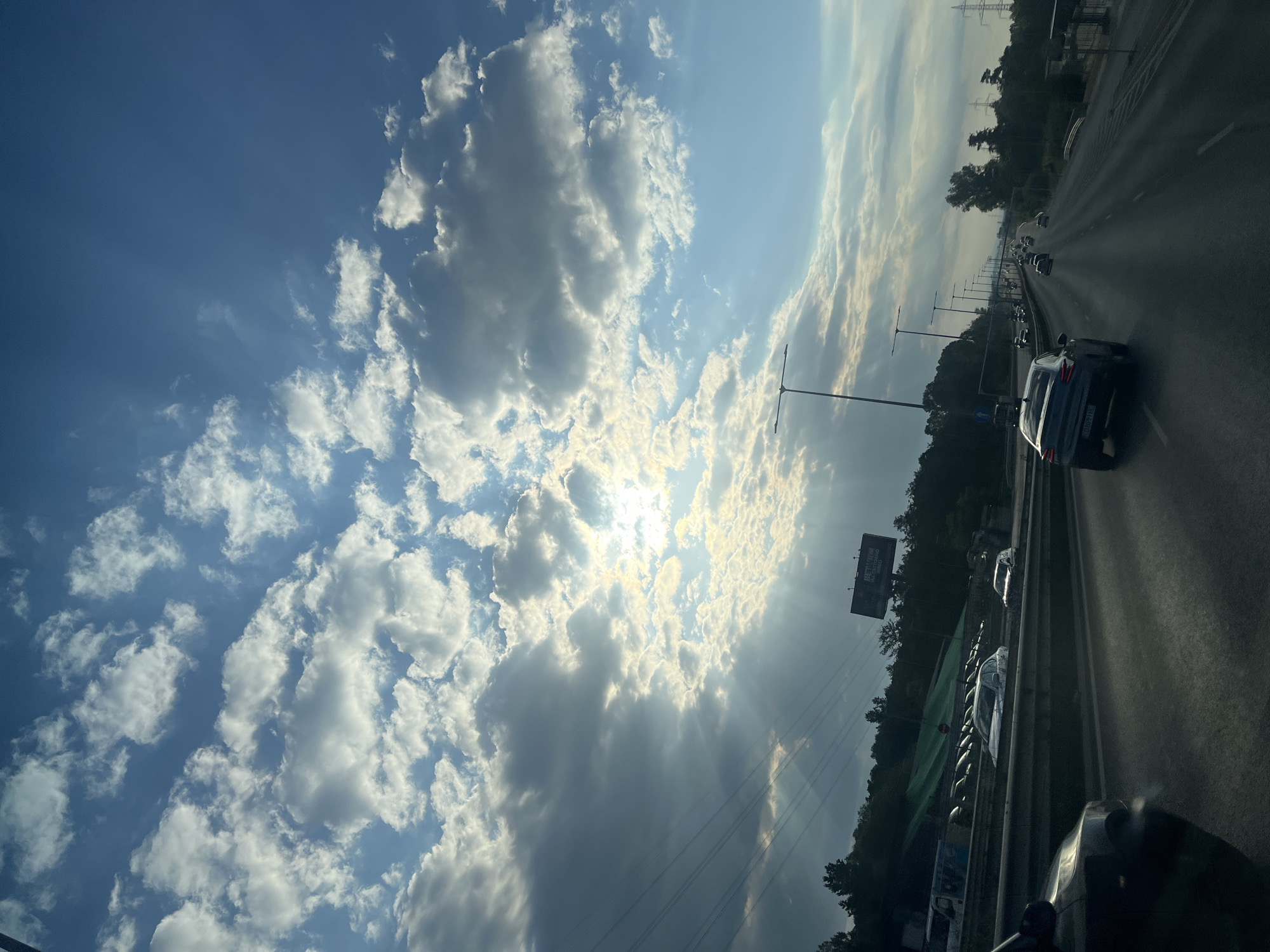 The image depicts a highway scene at dusk, with the sun breaking through dramatic clouds, casting rays of light across the sky. Several cars are traveling on the multi-lane road, including a dark-colored sedan in the foreground and a dark SUV in the middle lane. Trees and power lines line the sides of the highway, and a billboard is visible in the distance. The sky is a mixture of blue and grey, with fluffy white clouds. The overall impression is one of a dynamic and slightly moody sky over a typical urban highway. The location is the city of Unknown, Unknown.