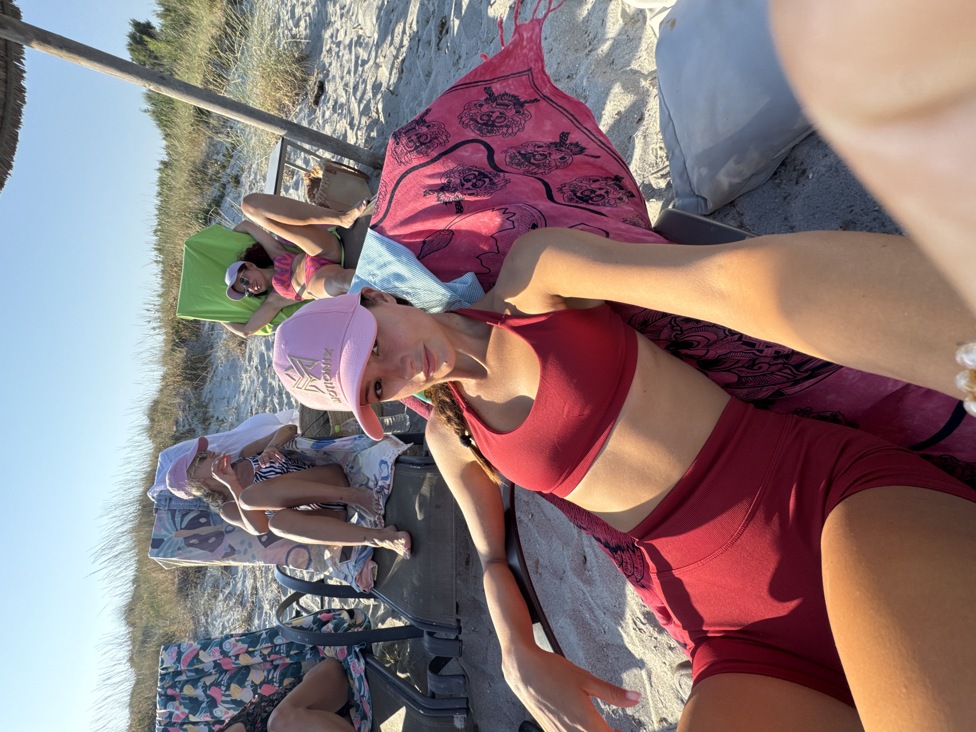 The image depicts a group of young women relaxing on a beach in Greece. The scene is set on a sandy beach with dry grass and vegetation visible in the background, suggesting a sunny and warm day. In the foreground, a woman wearing a red bikini and a pink baseball cap is lying on a beach chair, looking towards the camera. Another woman in a pink and black striped bikini is also visible lying on a chair to the left. Several beach chairs are arranged on the sand, some covered with colorful towels. A large pink towel with a black graphic pattern is spread out on one of the chairs. The time of day appears to be late afternoon or early evening, as indicated by the long shadows cast by the chairs and vegetation, and the warm, golden light. The overall mood is relaxed and casual, with the women enjoying a day at the beach. The baseball cap worn by the woman in the foreground has the word "MOTION" visible on it.
