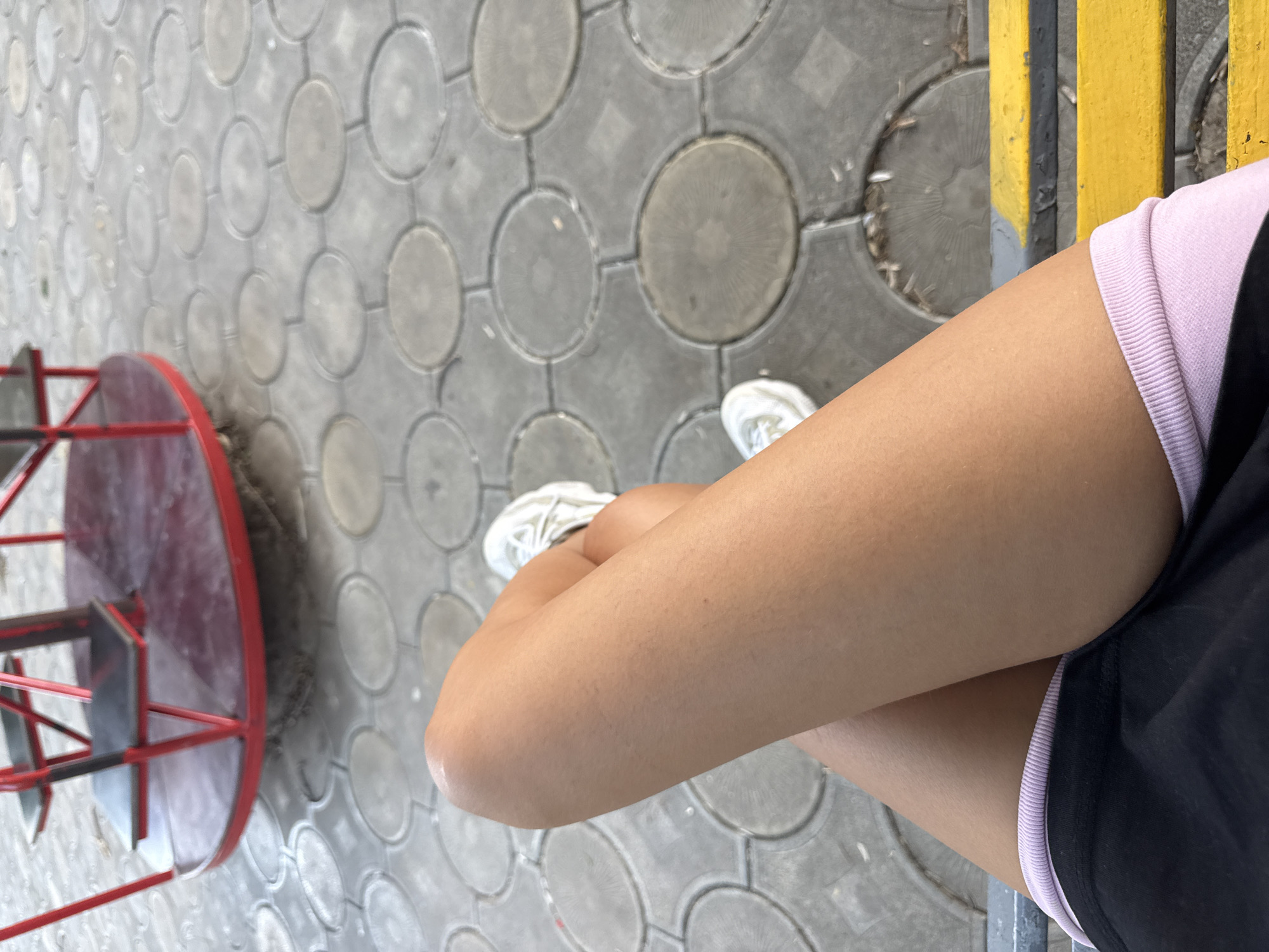 The image shows a close-up of a person's legs and feet, resting on a yellow bench. The person is wearing black shorts with a light pink trim and white sneakers with pink laces. The legs are tanned and appear to be smooth. The background is a paved area with circular patterns, possibly a playground, with a red and black merry-go-round visible in the upper left corner. The location is identified as Basarabeasca, Moldova.