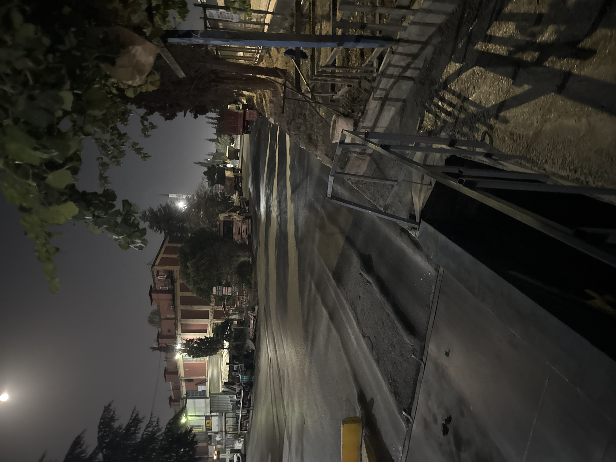The image depicts an outdoor scene at night, likely in Gebze, Turkey, characterized by dim lighting and shadows. A road occupies the foreground and midground, appearing wet, suggesting recent rain or a cleaning. Several vehicles, including trucks and what appears to be a car, are parked or moving along the road. Buildings, some with distinct architectural features like red facades and chimneys, are visible in the background. Trees and foliage are present throughout the scene, with some branches extending into the frame. In the distance, a minaret suggests the presence of a mosque. A metal railing structure runs along the right side of the image, alongside a dry, unpaved area. A light-colored bucket is visible near the railing. The overall atmosphere is that of an industrial or commercial area at night.