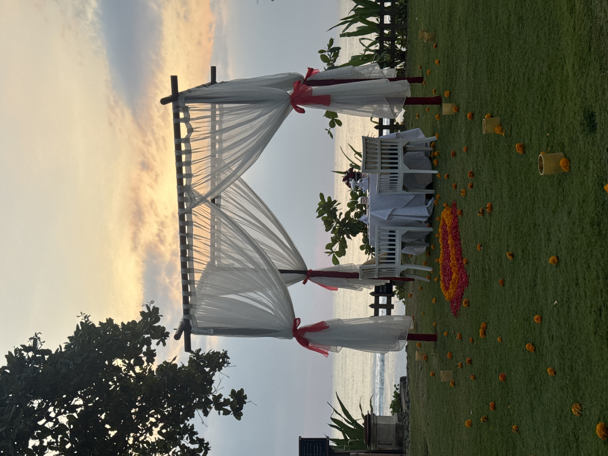 This is a wide shot of a romantic dinner setup on a grassy lawn by the ocean, likely in Kuta Selatan, Indonesia. The scene is set during sunset, with a soft, hazy sky in pastel colors.
Dominating the foreground is a white canopy tent, adorned with sheer white drapes tied back with red ribbons. Beneath the tent, a white tablecloth covers a small dining table set with two white chairs. The table is set for two, with plates, cutlery, and glassware visible. A bouquet of red flowers is placed in the center of the table.
The lawn around the table is decorated with scattered marigold petals and small yellow cylindrical lanterns. A heart-shaped arrangement of red and orange flower petals is laid out on the grass in front of the table.
In the background, the ocean stretches to the horizon, with gentle waves visible. A wooden fence separates the lawn from the beach. Lush green foliage lines the sides of the scene, framing the dining setup. The overall atmosphere is serene and intimate, suggesting a special occasion.