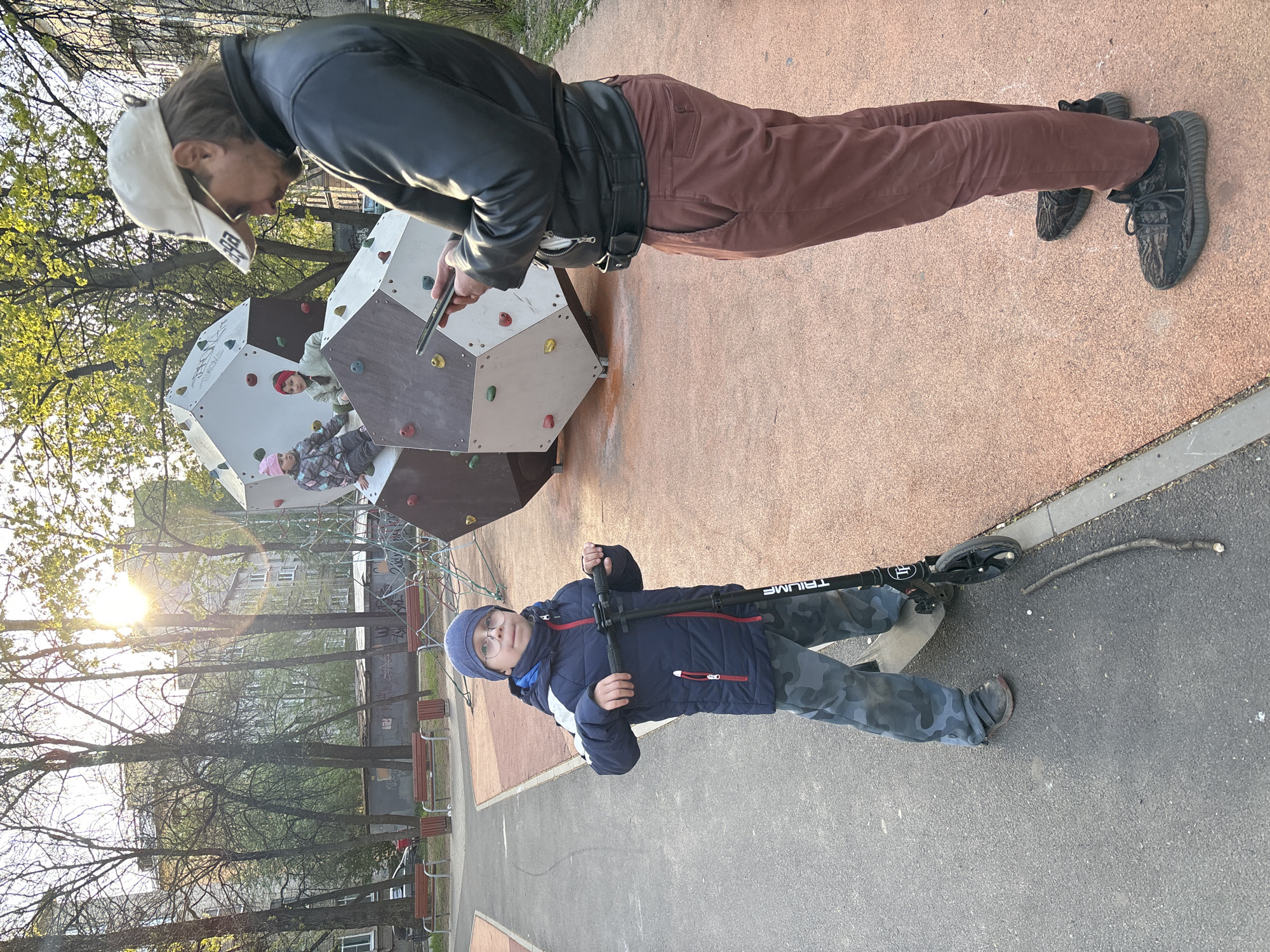 A man is photographing two children who are in a playground structure. The man is wearing a black leather jacket, brown pants, black sneakers, and a baseball cap. The boy on the ground is wearing a blue jacket, gray pants, and a blue hat and is holding a scooter. The playground structure is multifaceted and has climbing holds on it. The setting appears to be a park with benches and trees. The location may be Saint Petersburg, Russia.