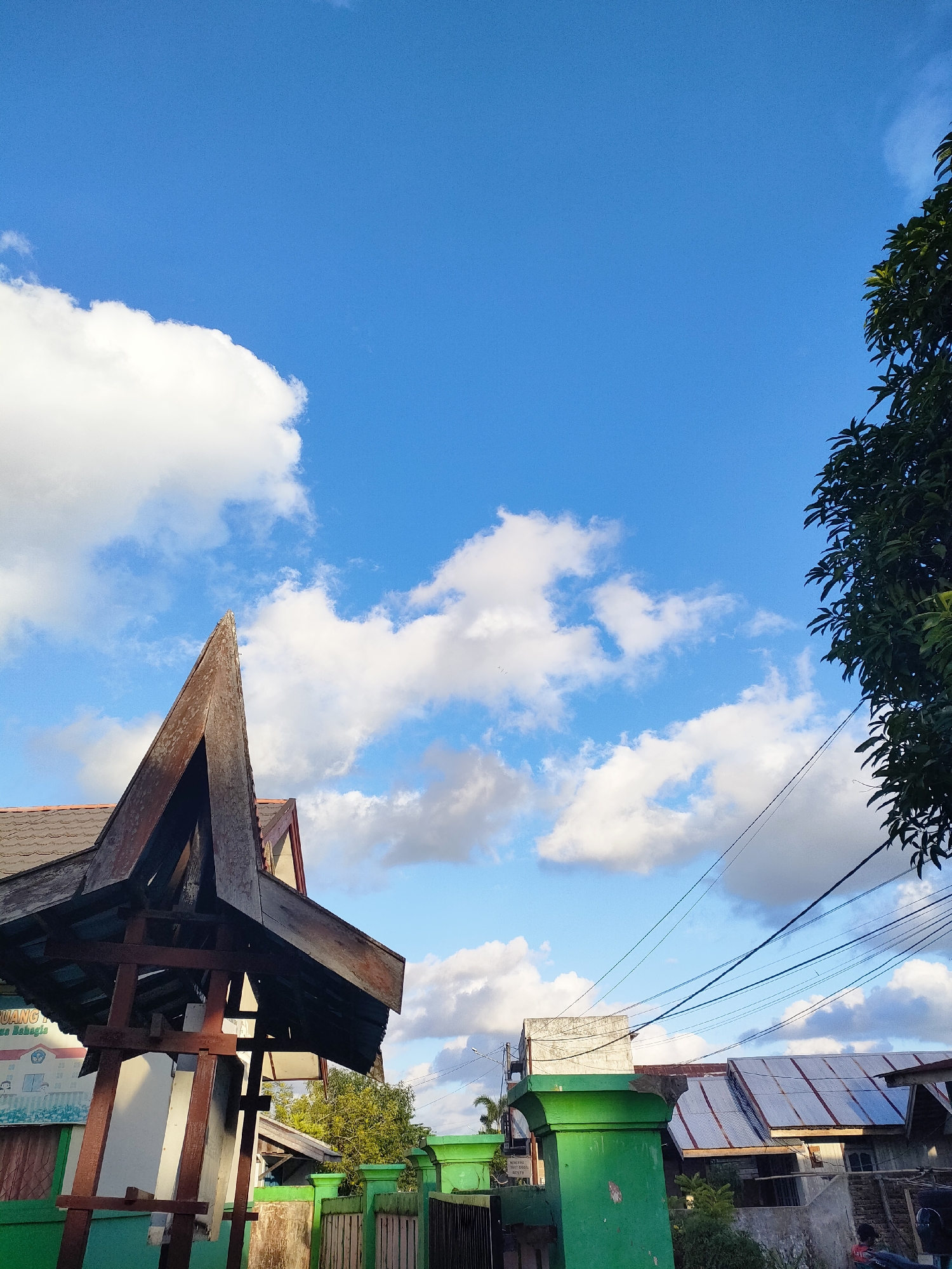The image depicts an outdoor scene under a bright blue sky with scattered white clouds. In the foreground, a portion of a wooden structure with a pointed roof is visible on the left, with a white wall and a sign behind it. A green fence and gate are also present. To the right, several houses with corrugated metal roofs and electrical wires running overhead are visible. A tree with green foliage is in the upper right corner. The scene appears to be in a residential or semi-urban area. No people are clearly identifiable in the image.
