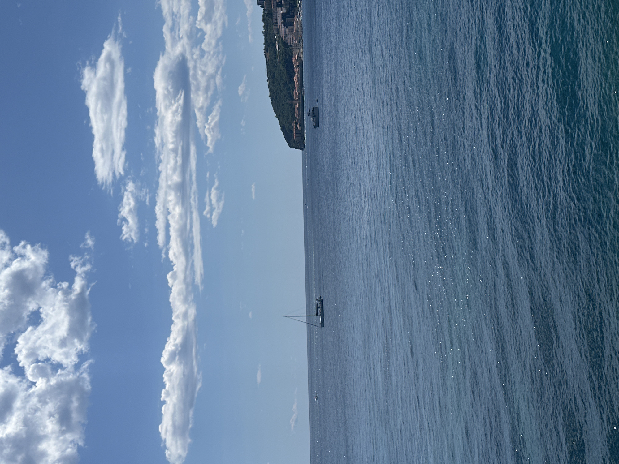 A wide shot captures a serene coastal scene in Budva, Montenegro, under a bright blue sky dotted with fluffy white clouds. The calm, deep blue sea occupies the majority of the frame, with gentle ripples reflecting the sunlight, creating a sparkling effect on the water's surface.
In the middle distance, a white sailboat with a single mast glides across the water. To the right, a small white boat is visible near the shore. The background features a lush, green, tree-covered peninsula that juts out into the sea. Along the coastline of the peninsula, a collection of buildings with red-tiled roofs and white walls are nestled, indicating a town or village. The architecture suggests a Mediterranean style. The overall impression is one of tranquility and natural beauty, with human presence evident in the boats and the coastal settlement.